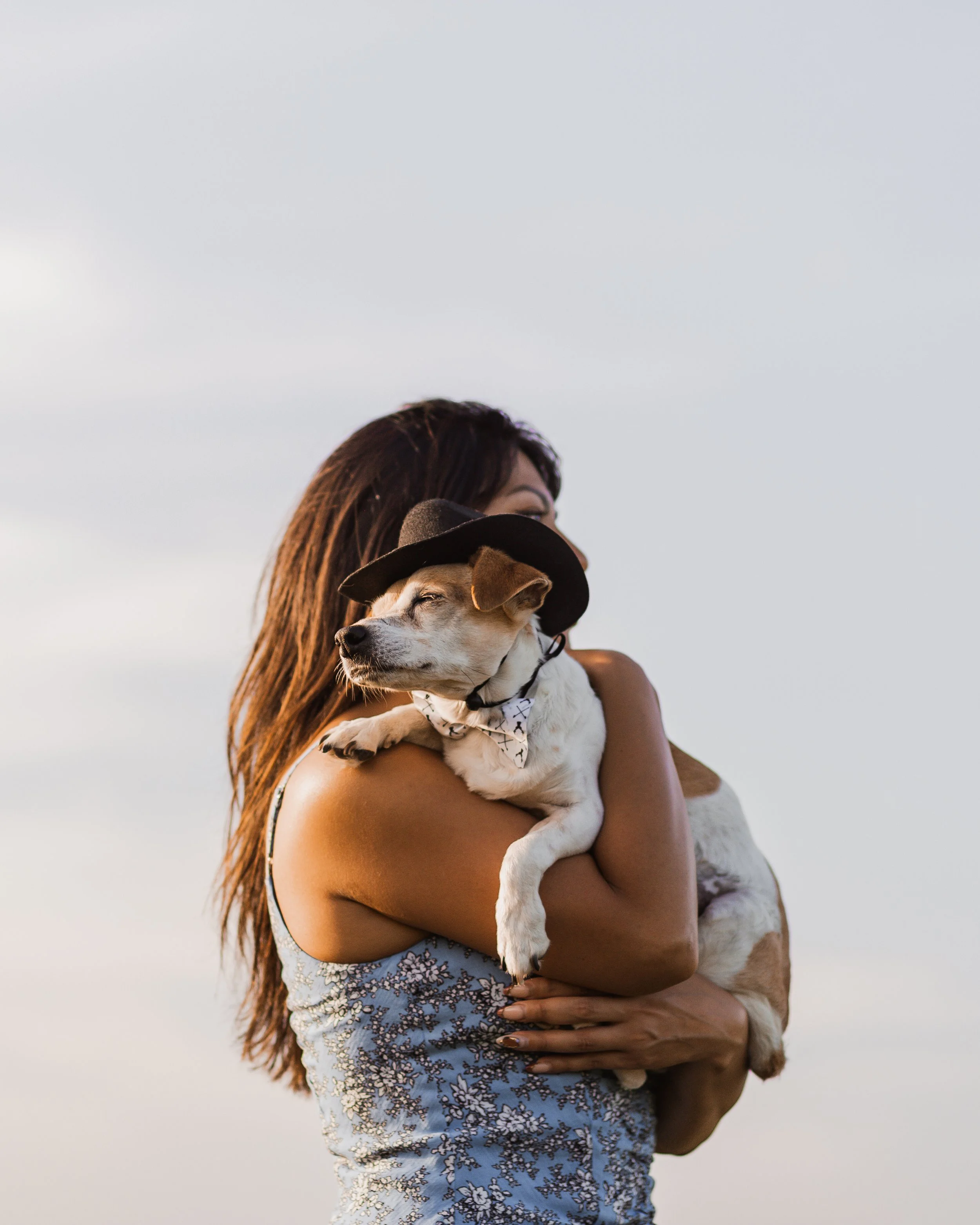 A woman with long brown hair holding a puppy with a bandana on its neck, against a cloudy sky background.