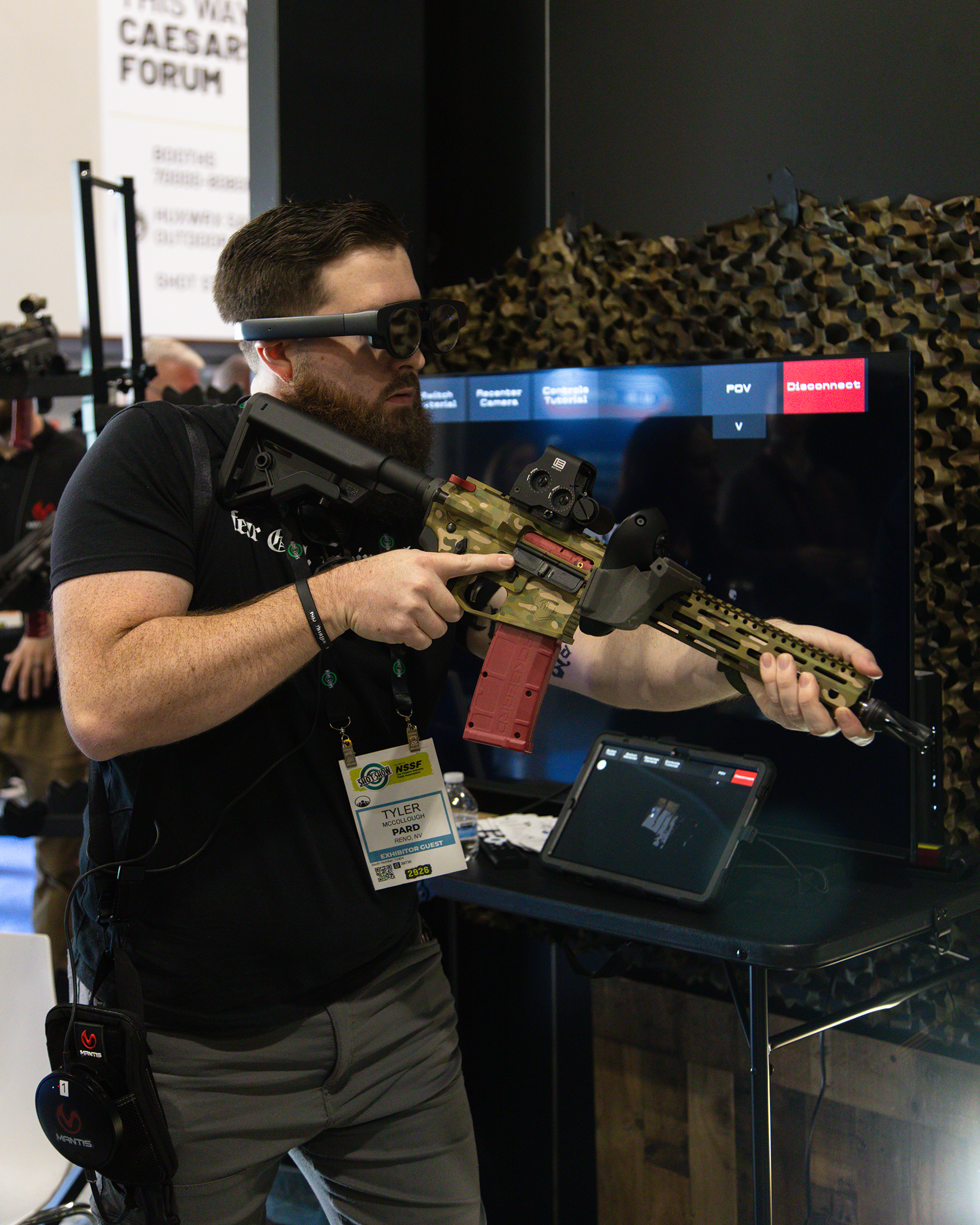 Man wearing augmented reality glasses aiming a camouflaged rifle at a screen with a red 'Disconnect' button at a tech exhibition.