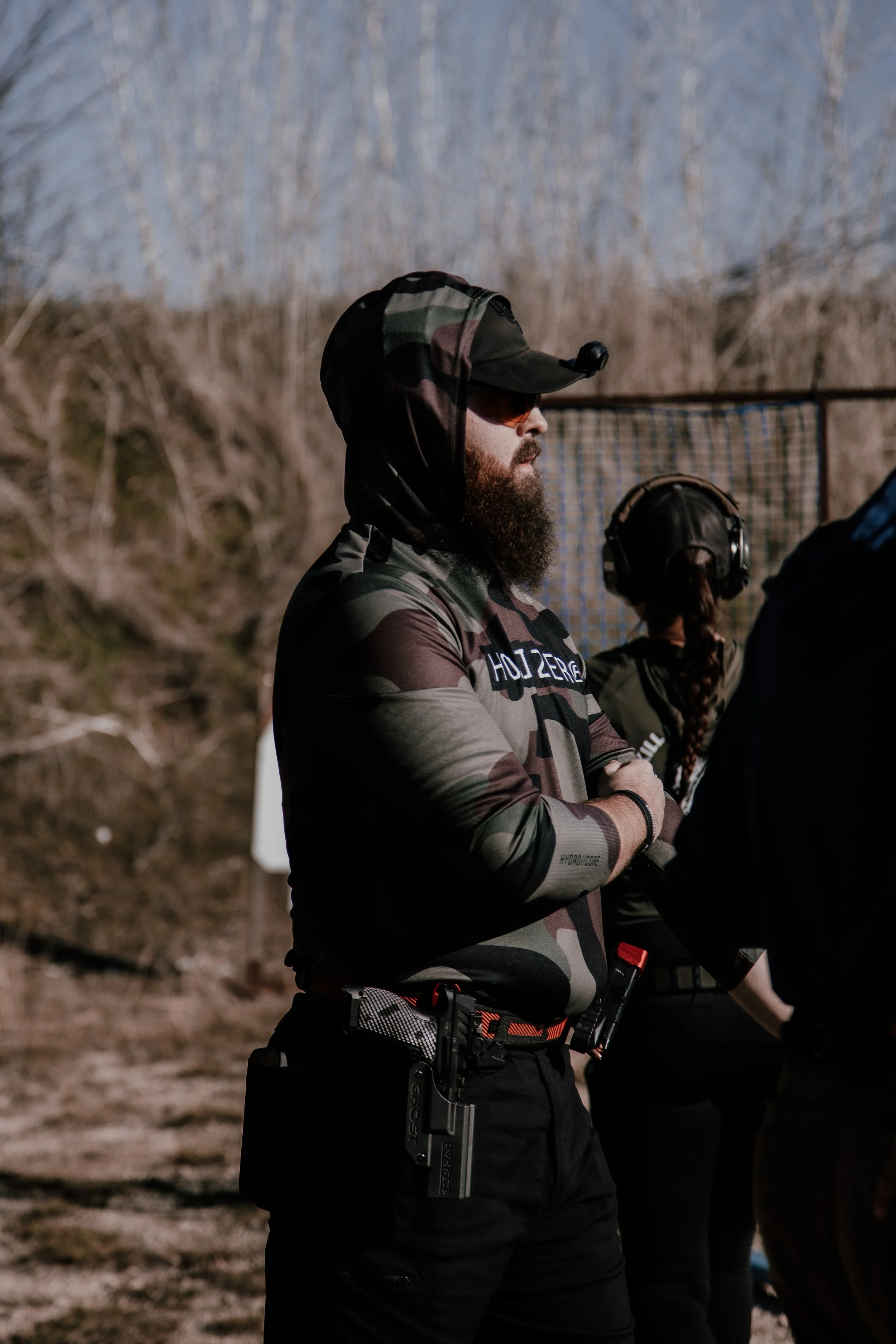 A man wearing camouflage clothing, a cap, and sunglasses standing outdoors in a wooded area with bare trees.