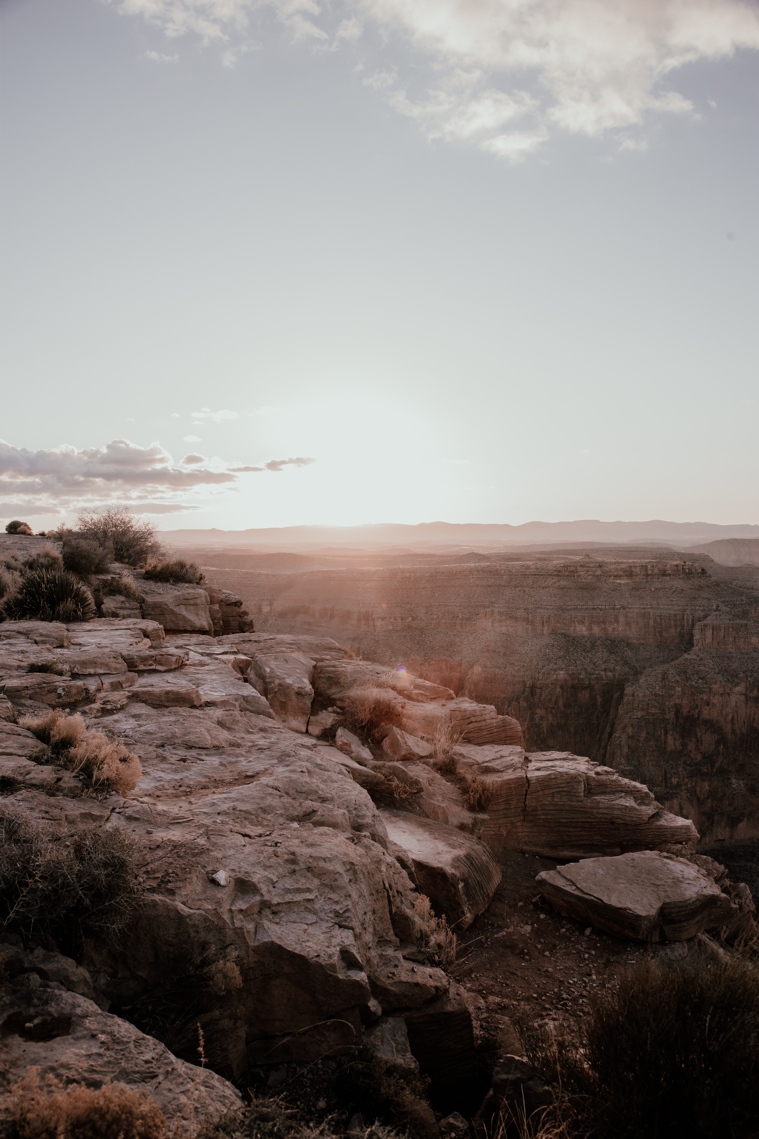 Sunset over a rocky canyon landscape with sparse desert vegetation in the foreground, clear sky with a few clouds.