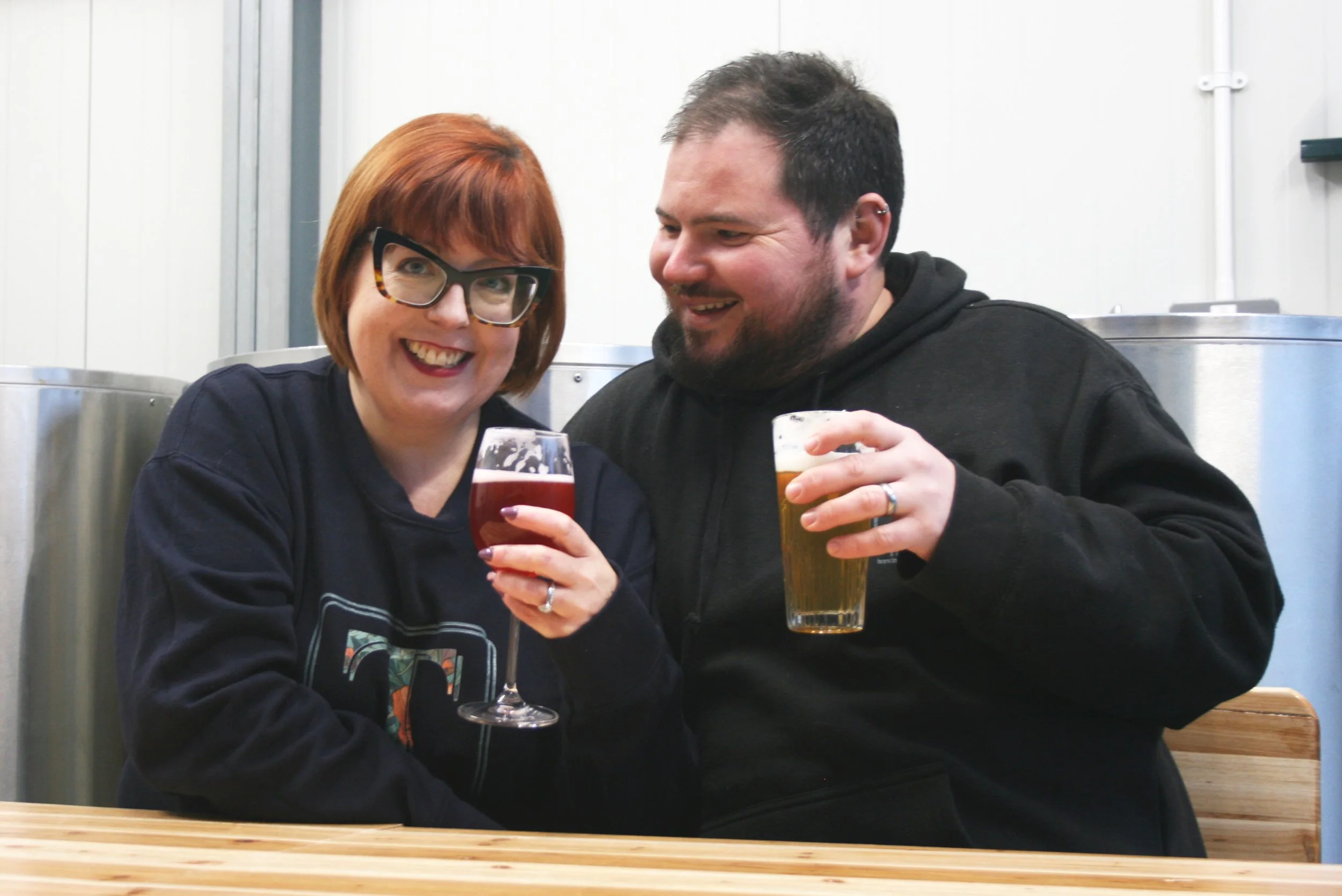 A smiling woman with glasses and red hair holding a glass of red wine, sitting next to a smiling man with dark hair and beard, holding a glass of beer, in a brewery or pub setting.