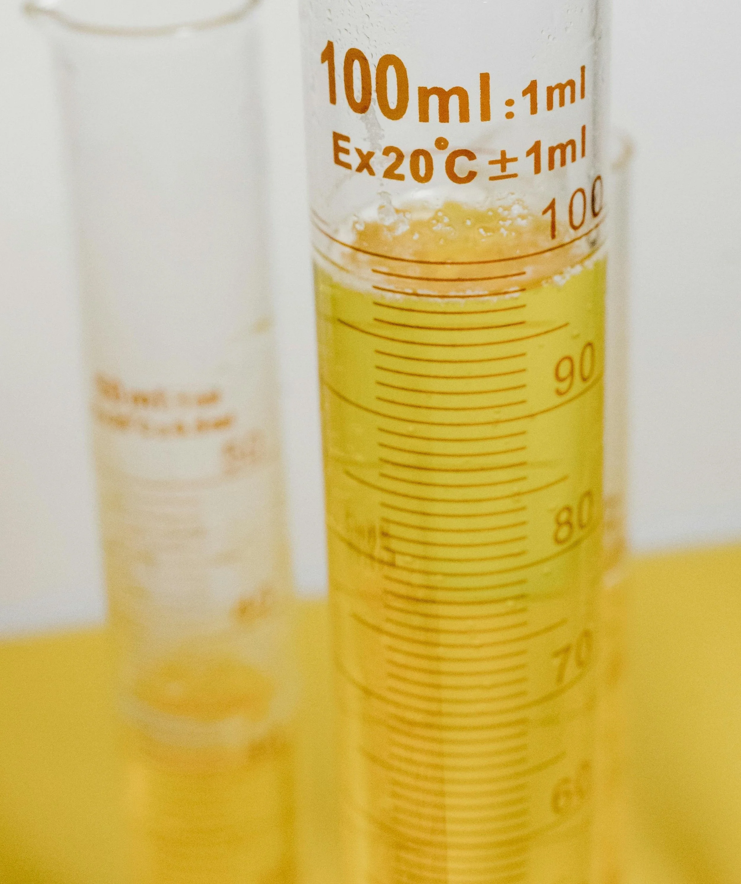 Close-up of a clear graduated cylinder with yellow liquid, marked with measurement lines in milliliters, placed on a yellow surface with another similar cylinder blurred in the background.