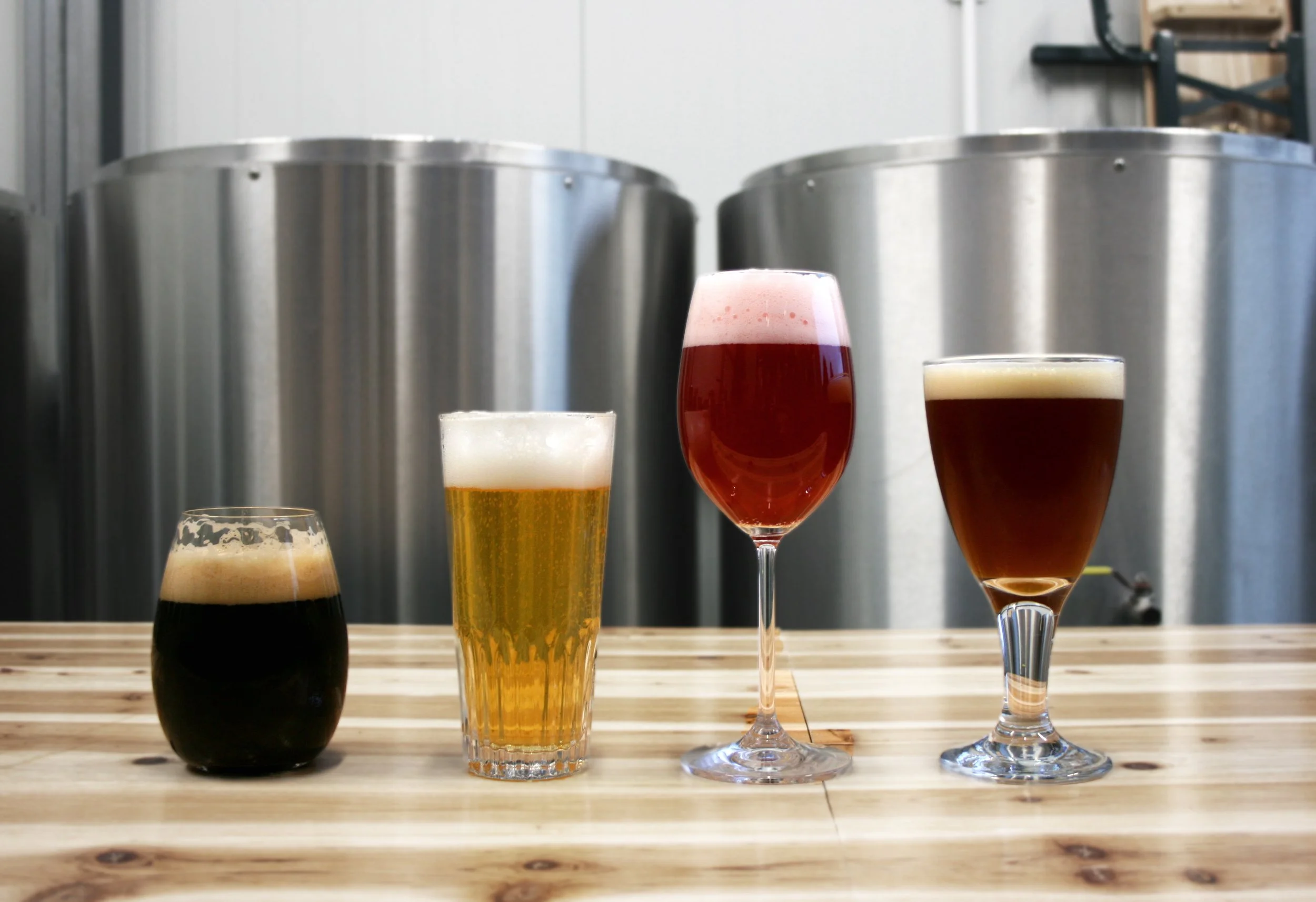 Four different glasses of beer lined up on a wooden surface with large metal brewing tanks in the background.
