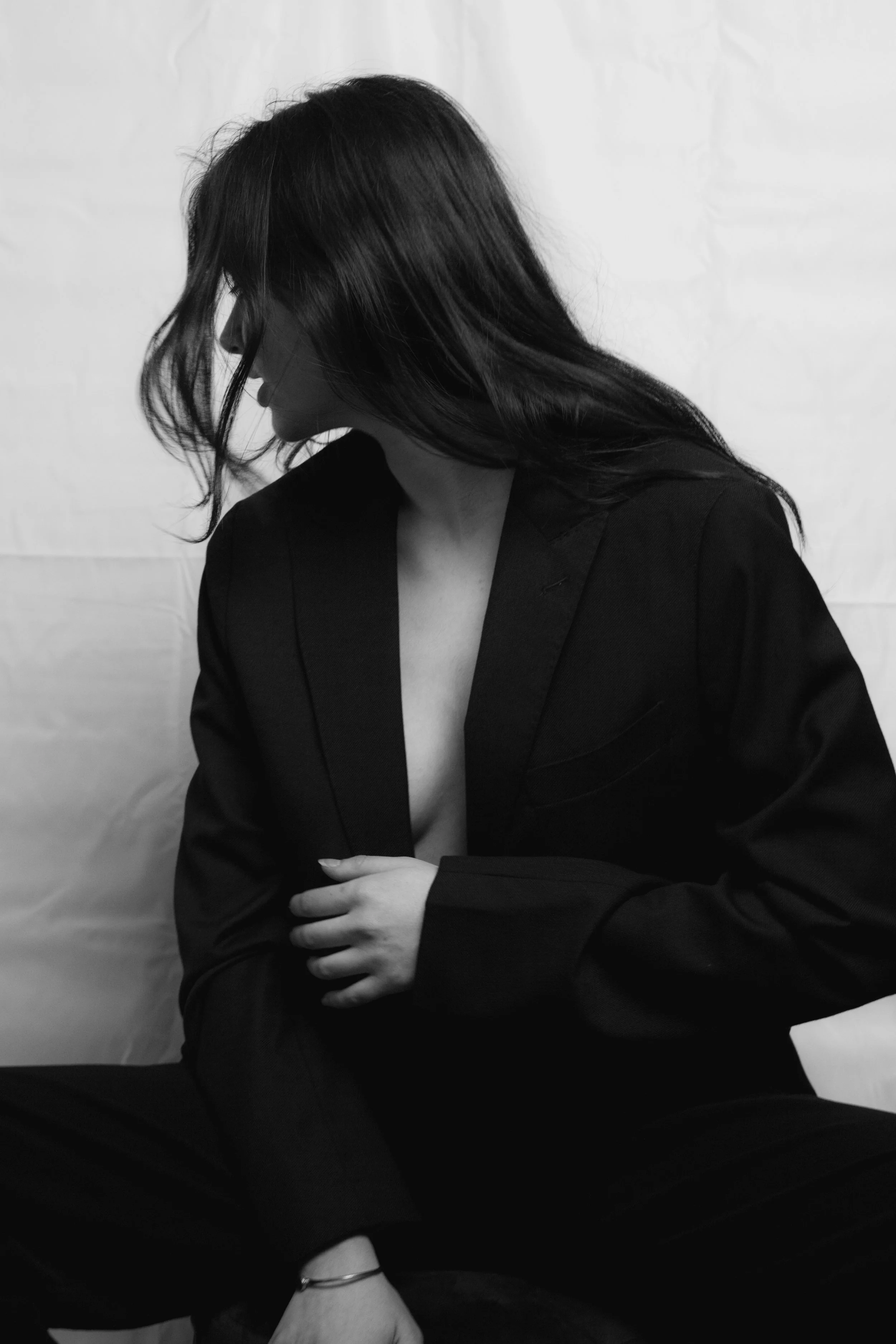 Black and white photo of a woman in a black blazer with her head tilted downward, hair covering part of her face, with her right arm resting on her lap and her left hand on her leg, in front of a plain, light background.
