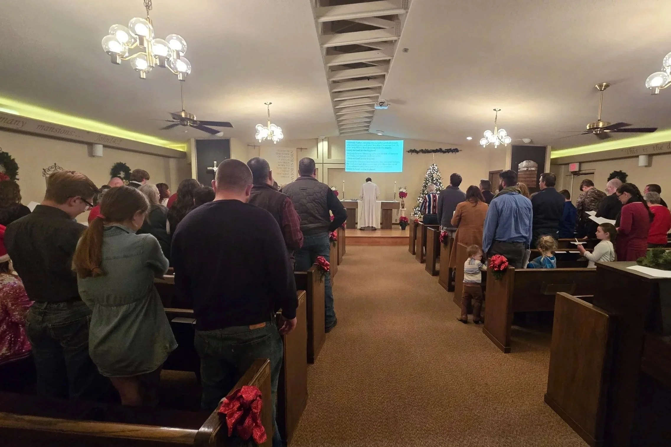 A congregation of people gathered in a church, standing and facing the altar, during a service. The church is decorated with Christmas ornaments, including a Christmas tree and poinsettias. The pastor is standing in front of the altar.