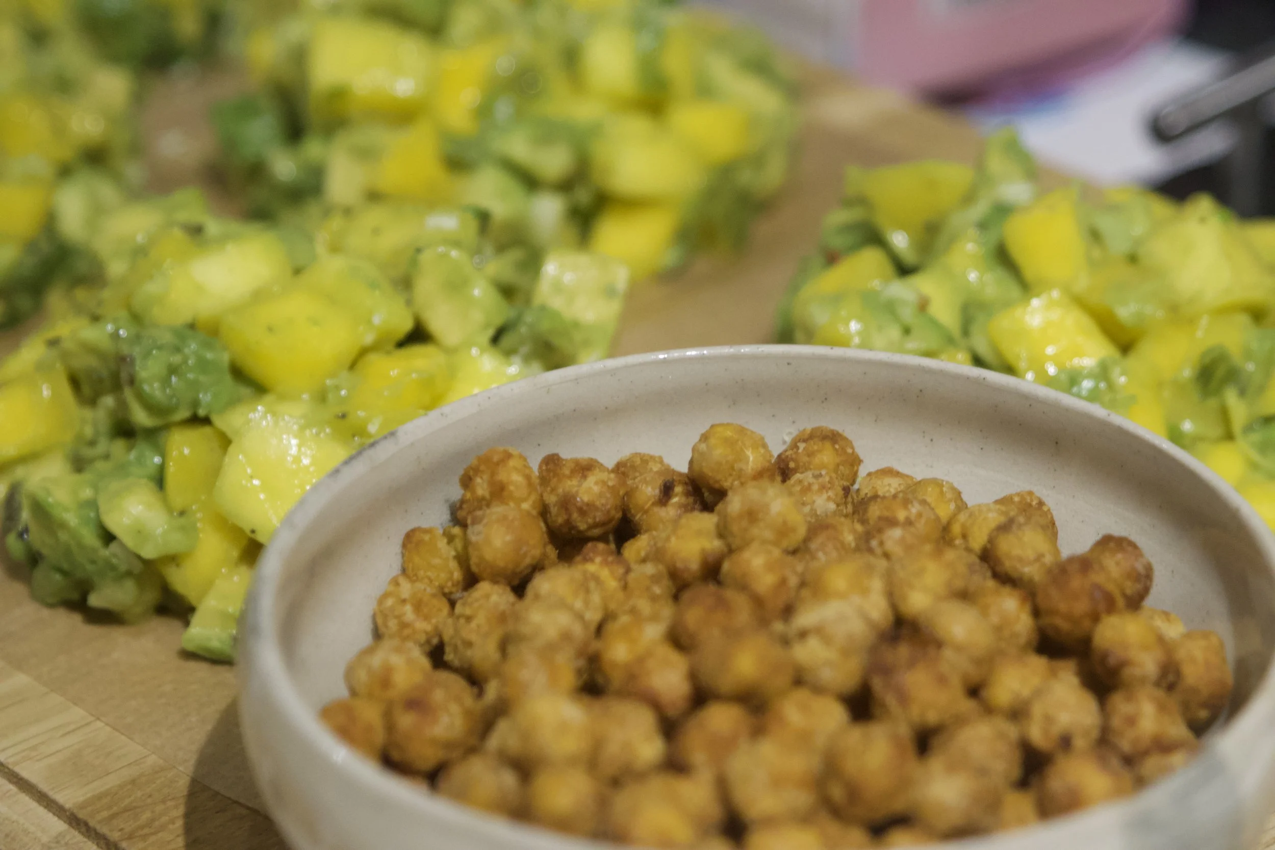 Close-up of a bowl of cooked chickpeas, with a blurred background of chopped mango and green avocado on a wooden surface.