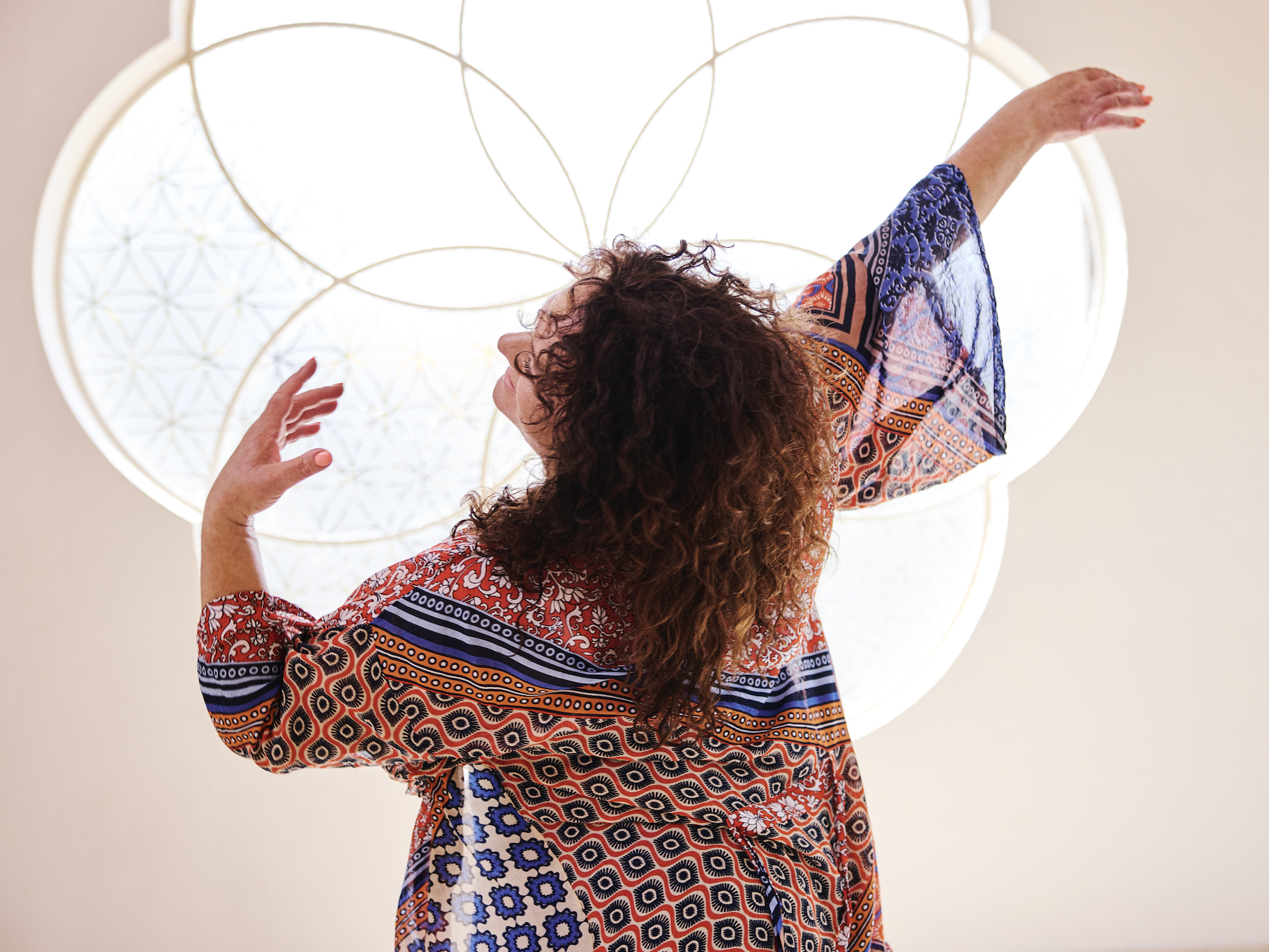 A woman with curly hair wearing a colorful patterned dress dancing or posing against a brightly lit, decorative circular ceiling fixture.