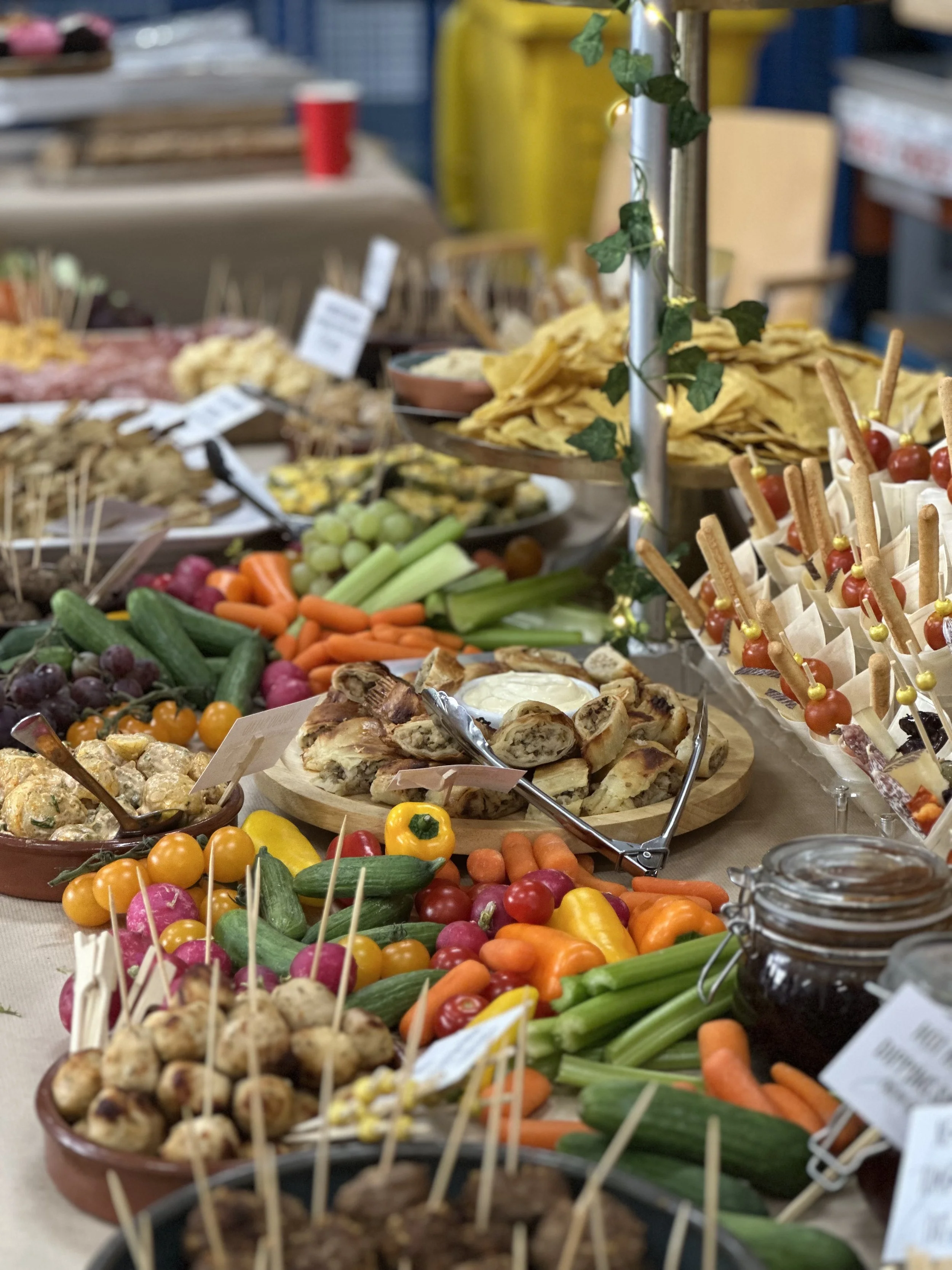 A buffet table filled with colorful fresh vegetables, fruits, and various appetizers, including cherry tomatoes, carrots, radishes, cucumbers, and small pastries, with some dishes garnished with herbs and decorative toothpicks.