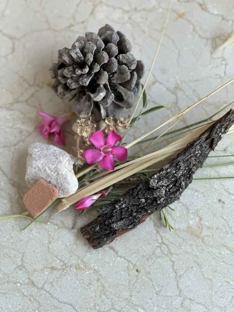 Arrangement of pinecone, pink flowers, dried grass, a small rock, a piece of bark, and a stick on a marble surface.