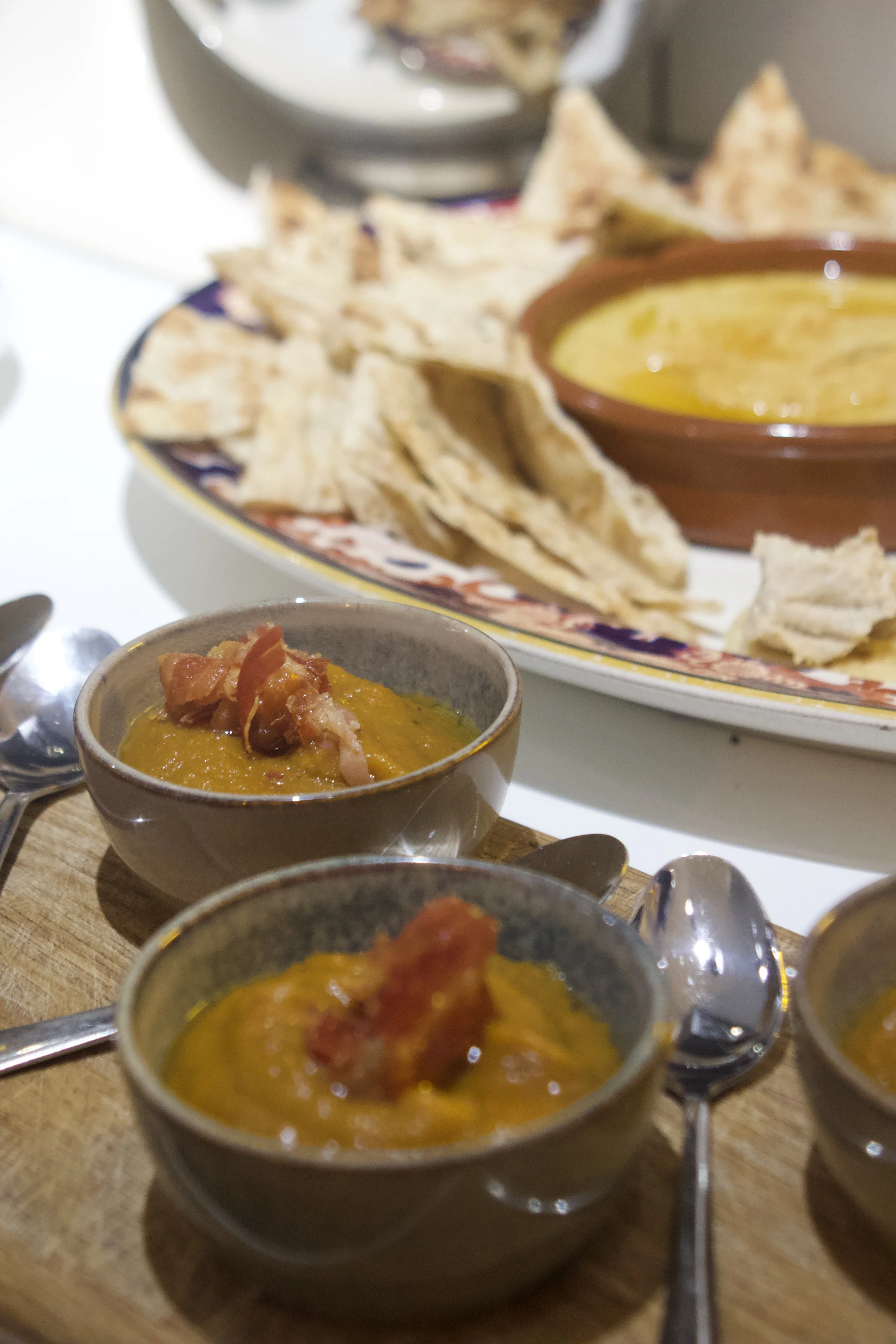 Close-up of small bowls of yellow lentil soup topped with bacon, small spoons, and a plate of pita chips with a small bowl of yellow sauce in the background.