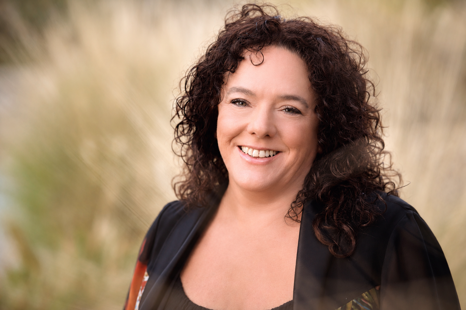 A woman with curly brown hair smiling outdoors, wearing a black top and jacket.