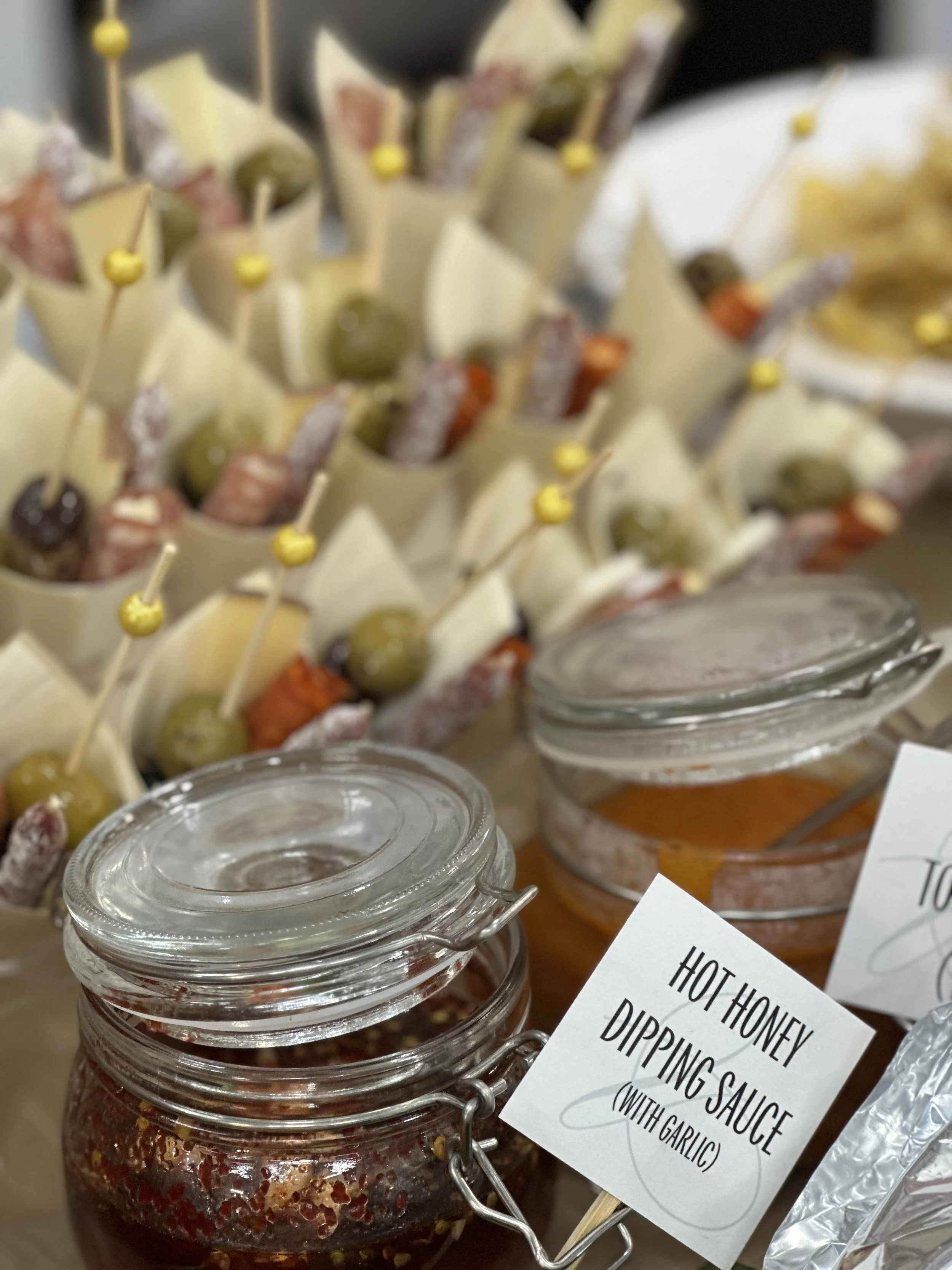 Jars of hot honey dipping sauce with green olives and skewered cheese, meats, and vegetables on display at a buffet or food station.