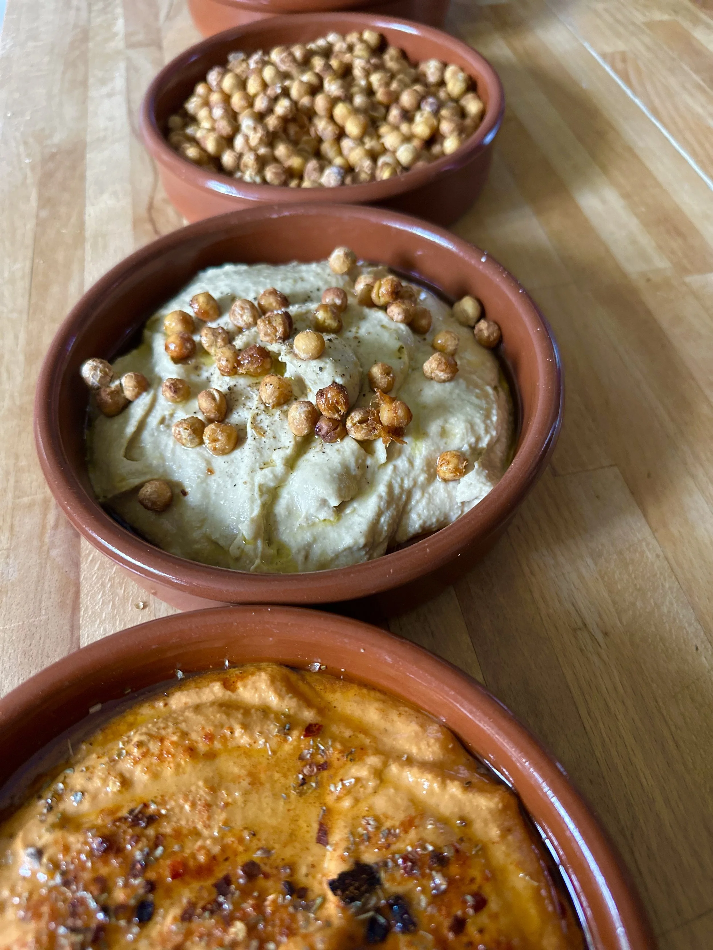 Three terracotta bowls filled with different types of food, focusing on an appetizer dish with hummus topped with chickpeas, and another dish with a hearty stew or sauce.