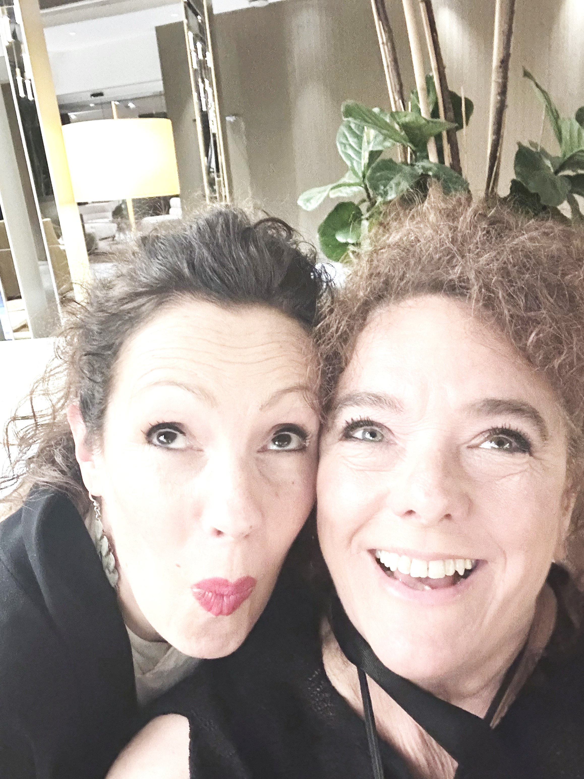 Two women smiling and posing closely together for a selfie in an indoor setting, with one woman having curly hair and the other with lighter hair. In the background, there is a potted plant and a mirror reflecting a brightly lit room.