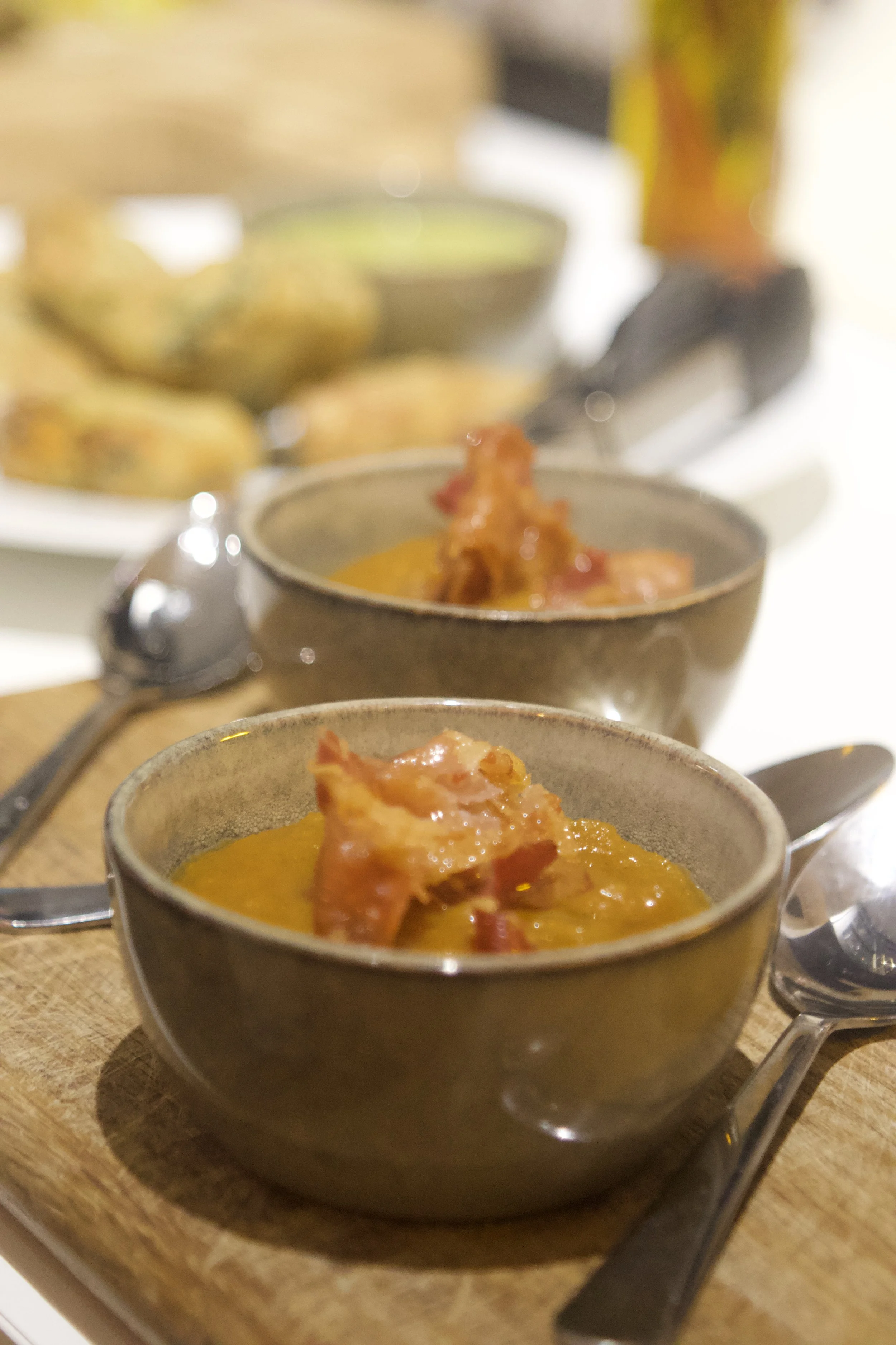 Close-up of a cup of yellow curry with meat, with a spoon inside, on a wooden table. In the background, there are other similar cups with curry, a bowl of snacks, and a blurred drink bottle.