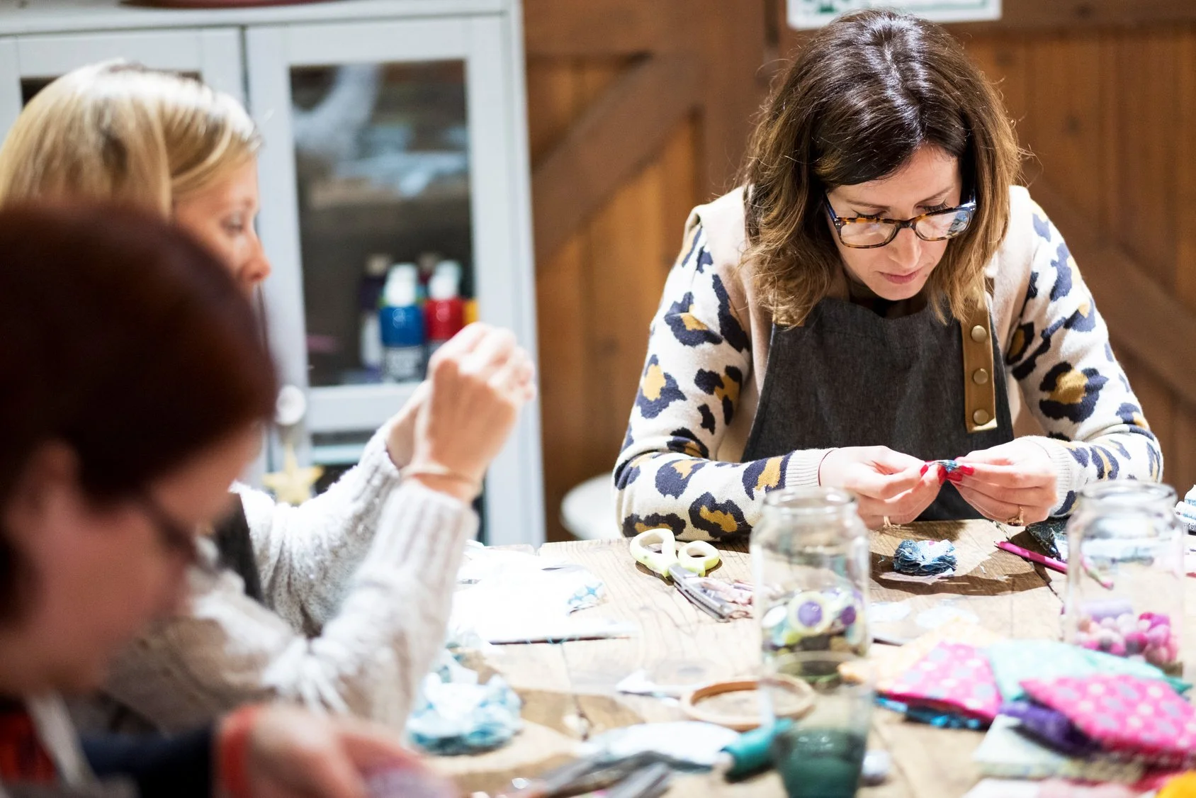 Women gathered around a table engaging in a craft activity, with various craft supplies and jars of decorative items on the table.