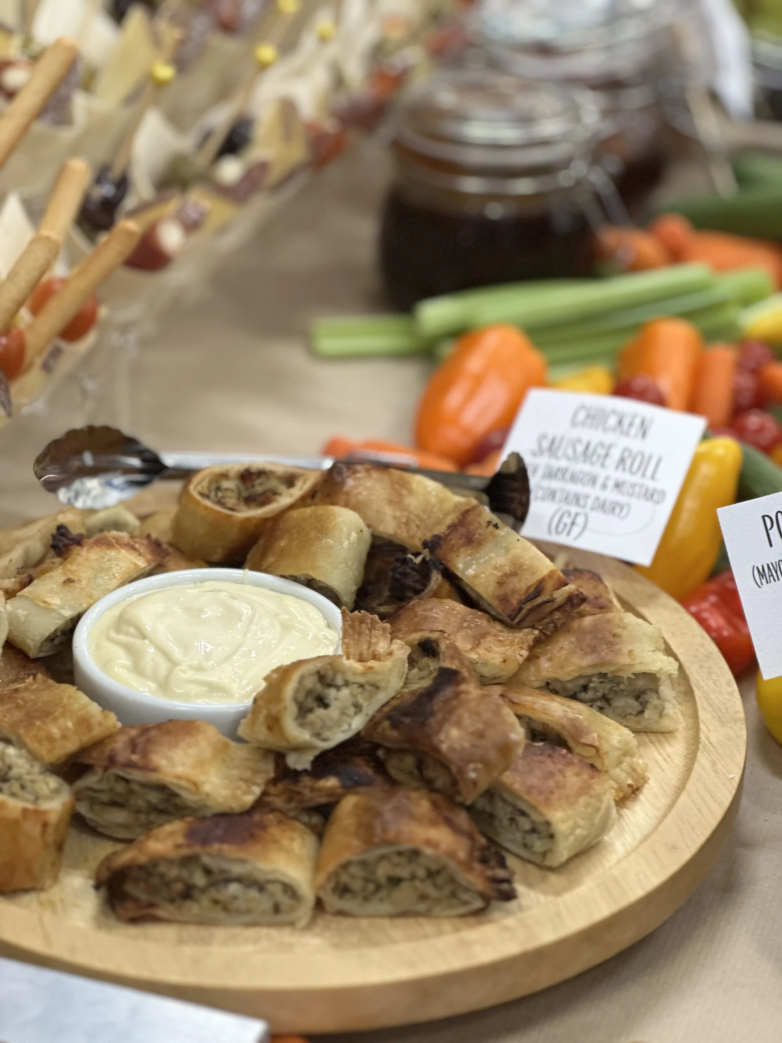 A wooden platter filled with  Homemade chicken Rolls with a side of creamy dipping sauce, surrounded by fresh vegetables and jars of jam in the background.