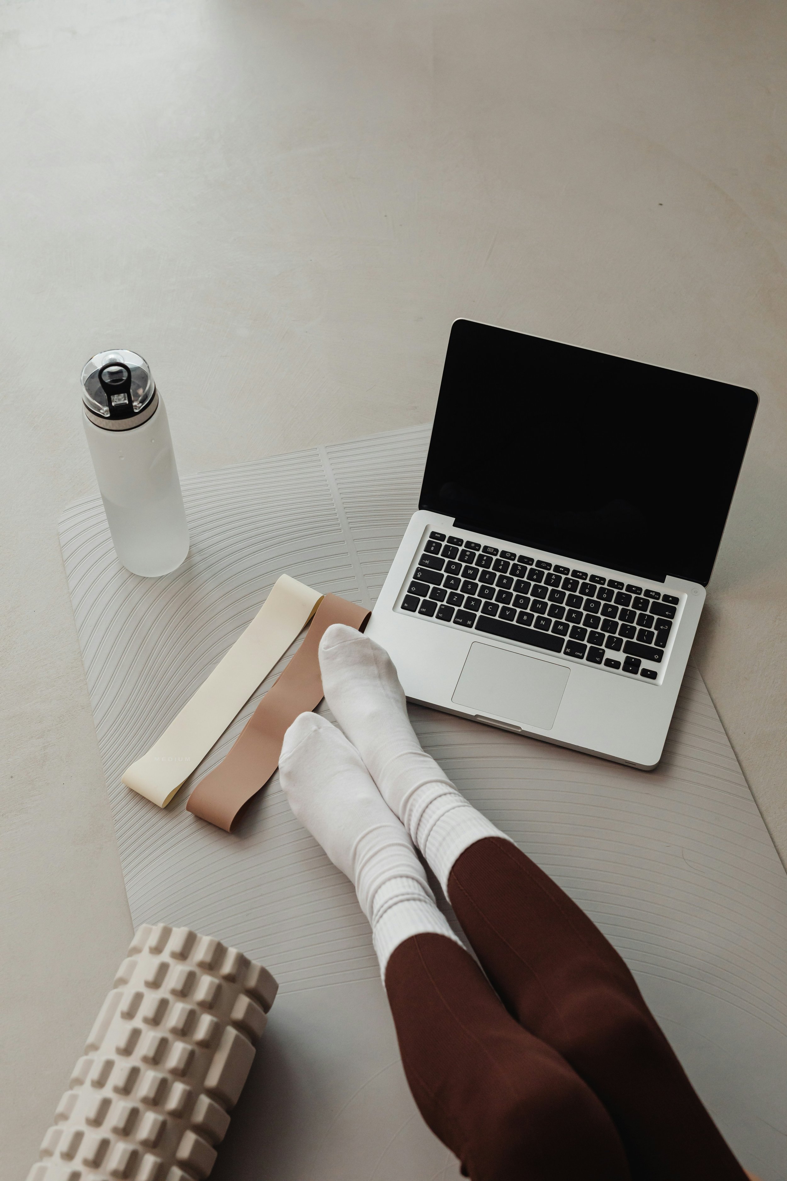 Person lying on a mat with legs extended, wearing white socks and brown pants, using a laptop, with a water bottle and color swatches nearby.