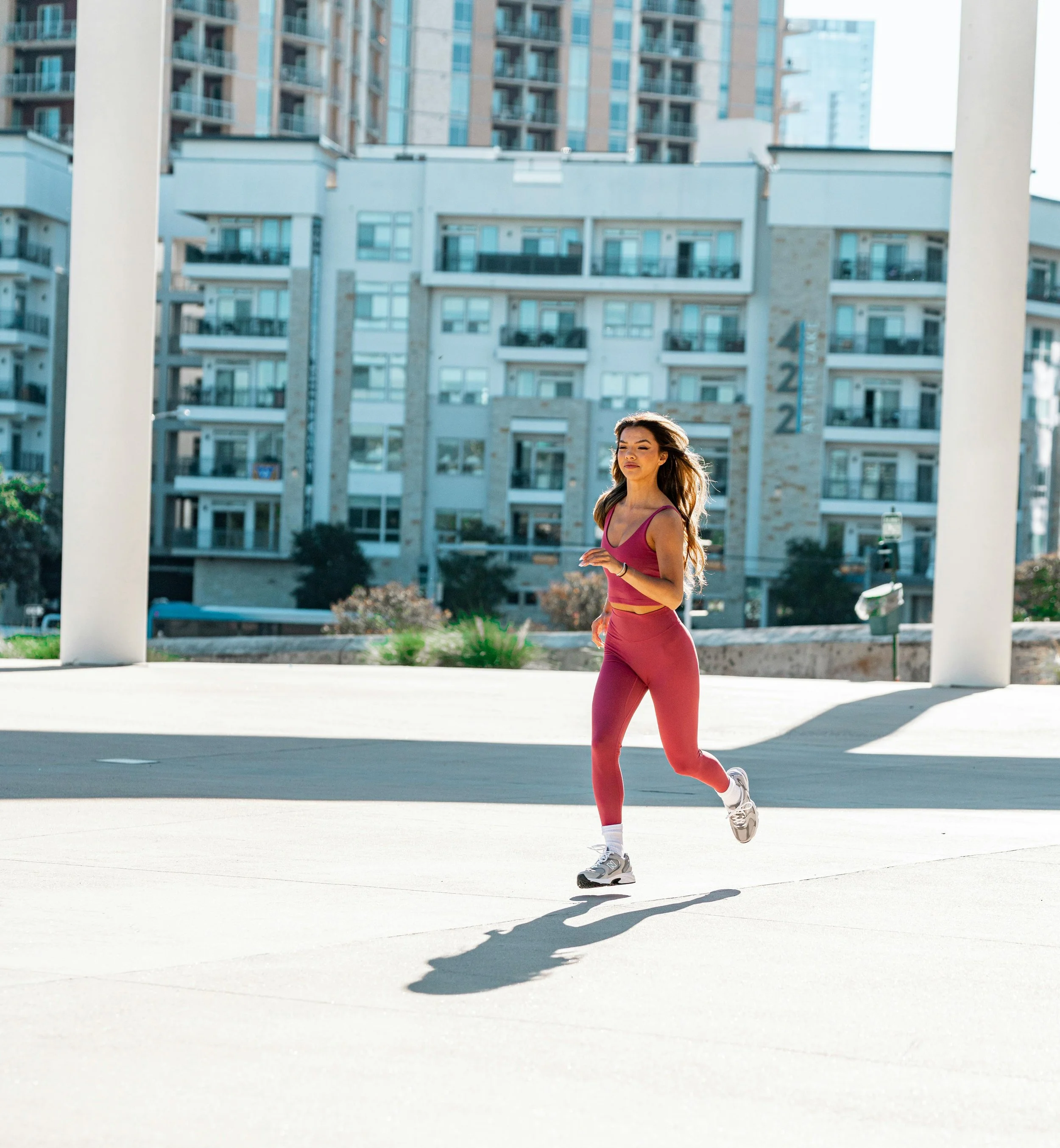 A woman running outdoors in a city setting, wearing a pink athletic outfit with buildings in the background.