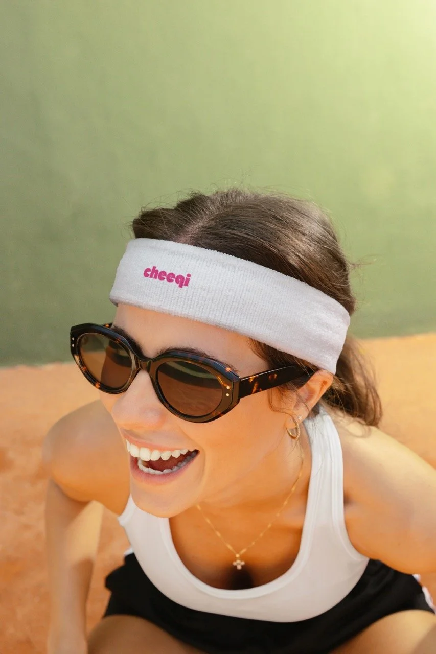 A woman smiling wearing sunglasses, a white headband with pink text, and a white tank top, sitting against a green wall.