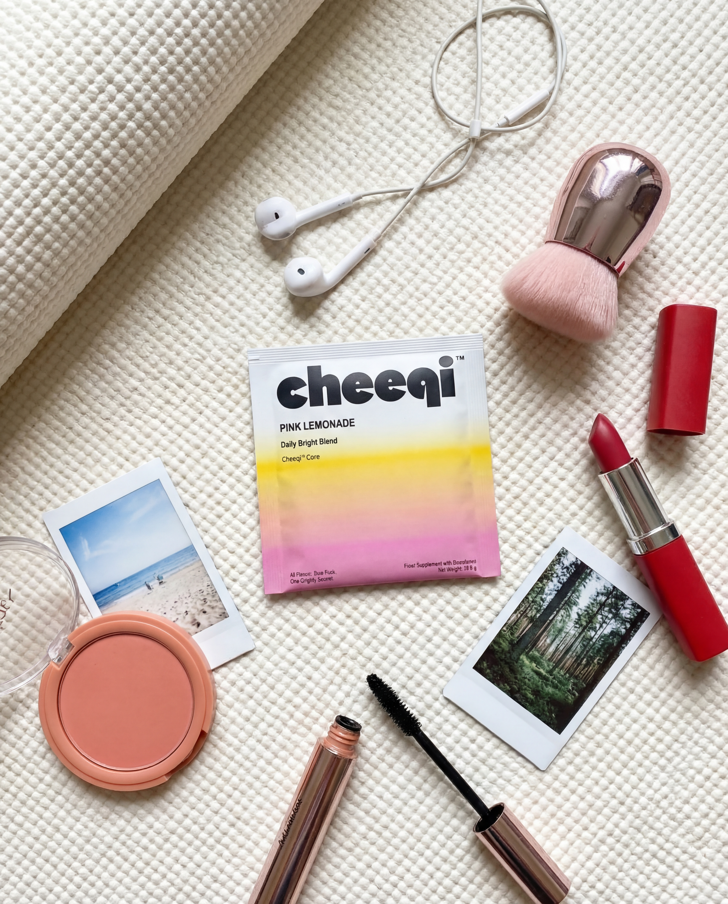 Flat lay of beauty and travel items on a white textured surface, including a pair of white earphones, a pink and rose gold makeup brush, a red lipstick, a pink blush compact, a tube of mascara, two small photo prints of nature scene and beach, and a packet of Cheeqi Pink Lemonade tea.