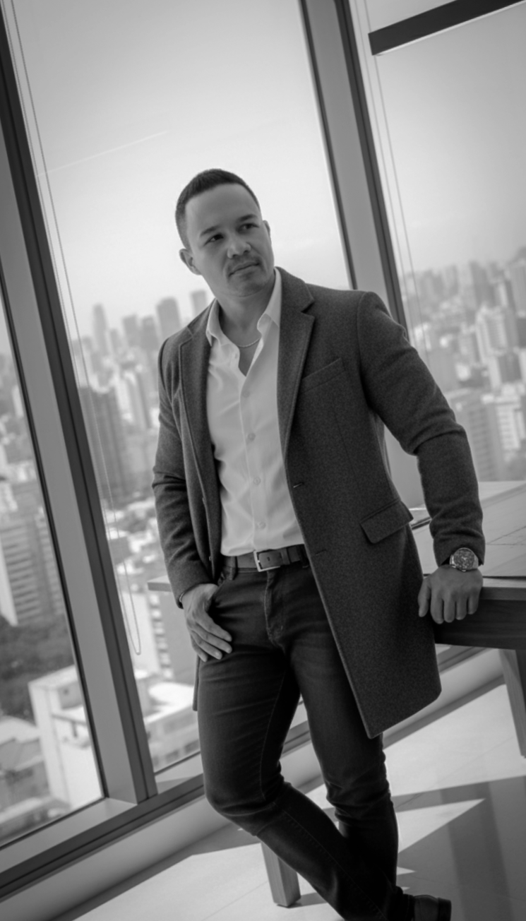 Hombre con traje y camisa blanca, de pie junto a una ventana con vista a la ciudad, en un entorno moderno y de oficina.