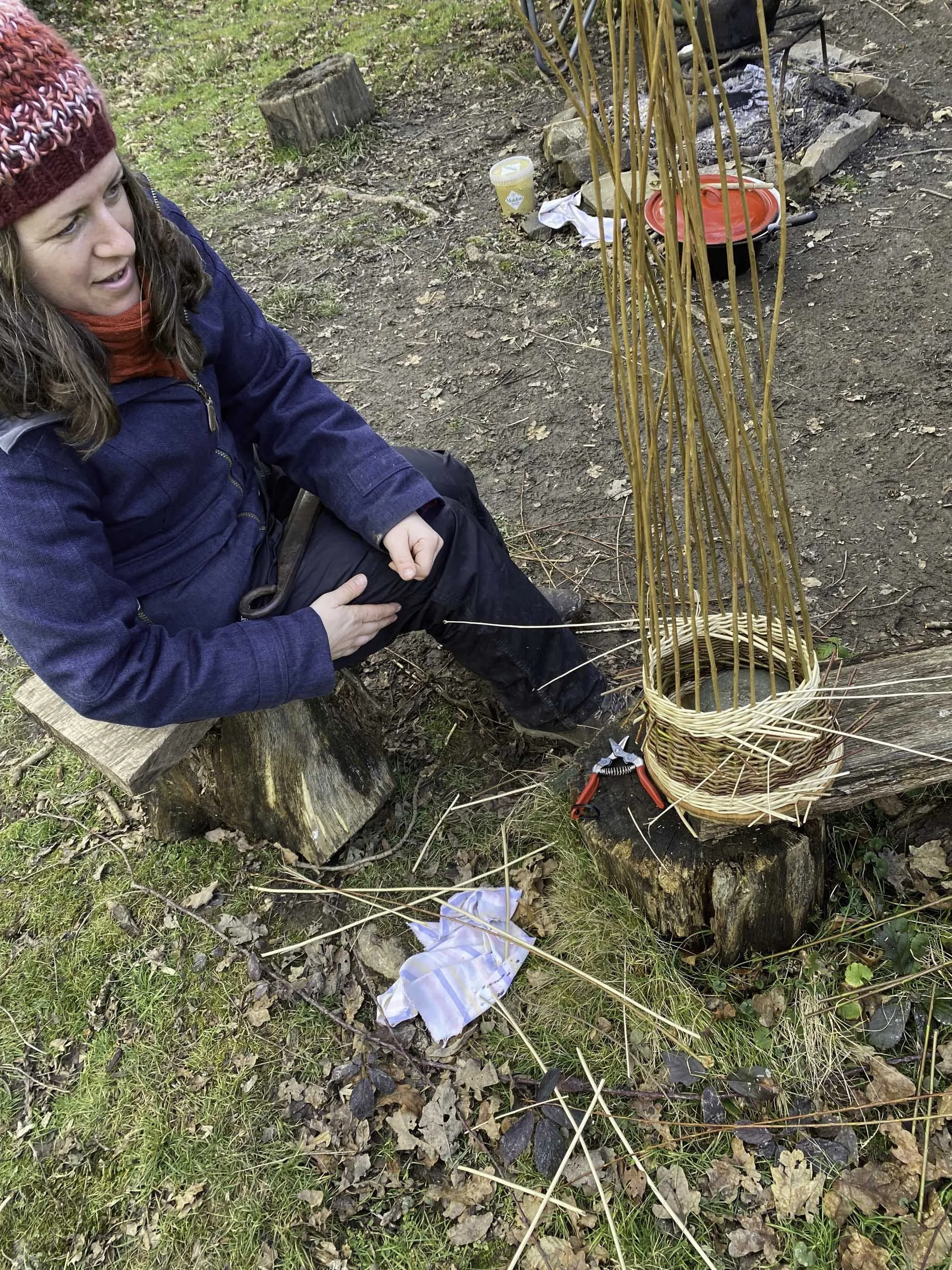 A woman in outdoor clothing, including a red knit hat and a blue jacket, sitting on a tree stump near a small outdoor fire pit with firewood and sticks, surrounded by natural ground with leaves and grass.