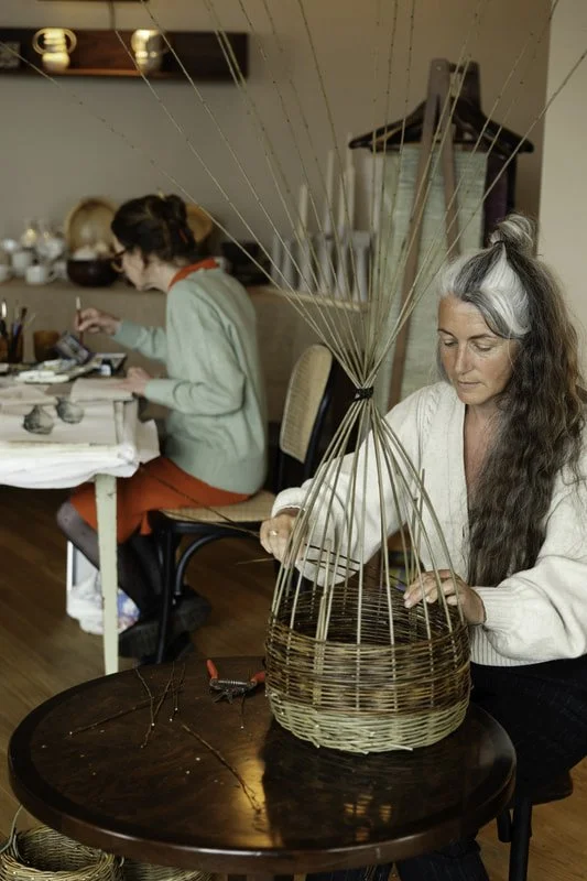 A woman weaving a basket at a table with another person working in the background.