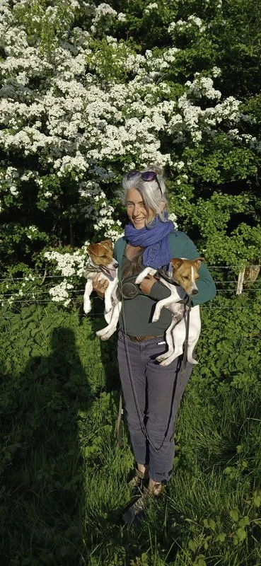 A smiling woman with gray hair wearing sunglasses, a teal shirt, and blue scarf, holding two small dogs, one in each arm, outdoors with green foliage and white flowering bushes in the background.