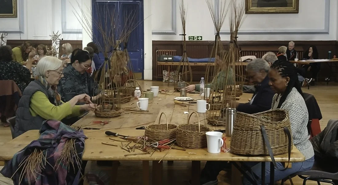 People sitting around a large wooden table, weaving baskets with natural materials inside a room with white walls and blue doors.