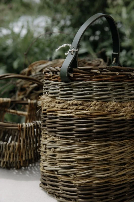 Close-up of a woven wicker basket with a metal handle, outdoors with green foliage in the background.