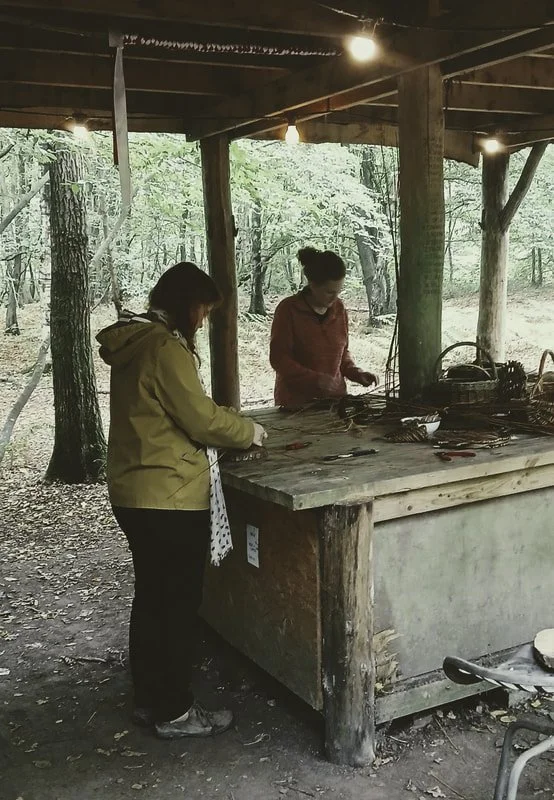 Two women are working at a rustic wooden outdoor workbench in a wooded area, possibly crafting or repairing items, under a shed with string lights.
