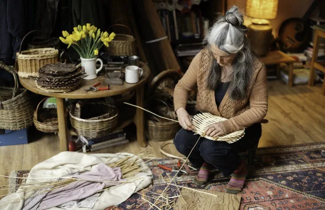 An elderly woman with gray hair in a bun, wearing a brown sweater and colorful socks, is sitting on a rug and weaving strands of straw or similar material in a cozy room with wooden walls and shelves. A table behind her holds a bouquet of yellow daffodils, a stack of dark chocolate cakes, mugs, and various baskets.