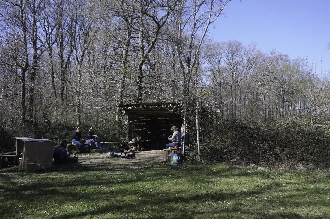 People sitting around a campfire near a wooded area with leafless trees, some under a small shelter, on a sunny day.