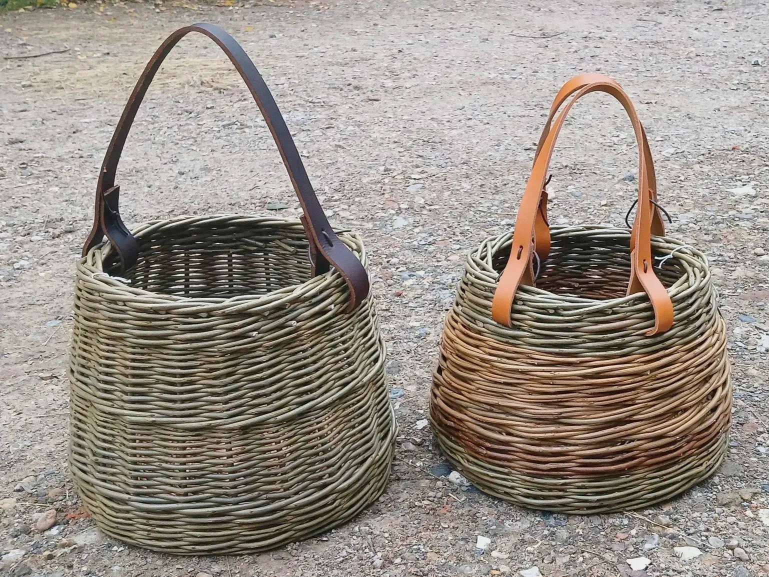 Two woven wicker baskets with leather handles, placed on a gravelly outdoor ground.