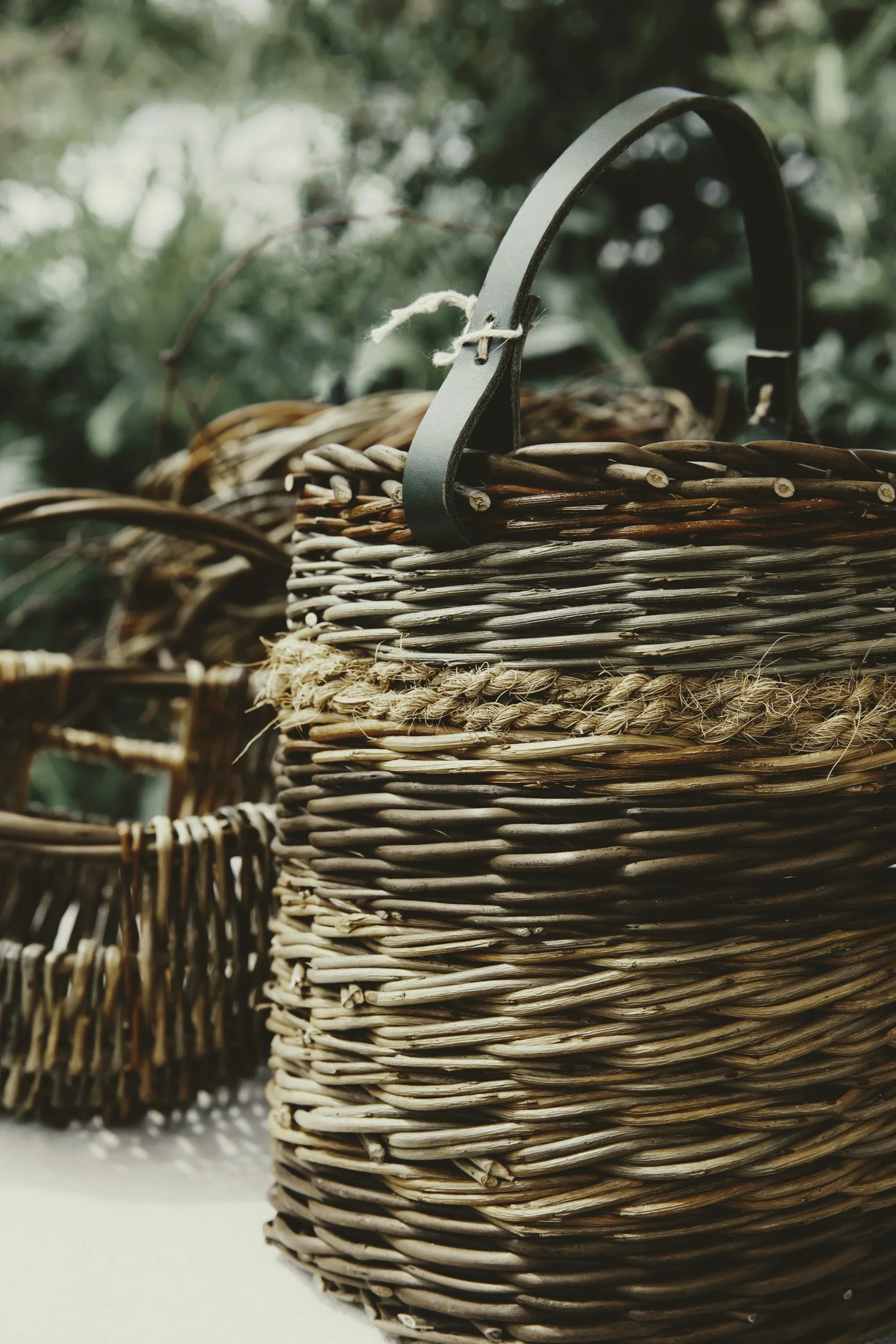 Close-up of woven baskets with a green metal handle, set outdoors with green blurred foliage in the background.