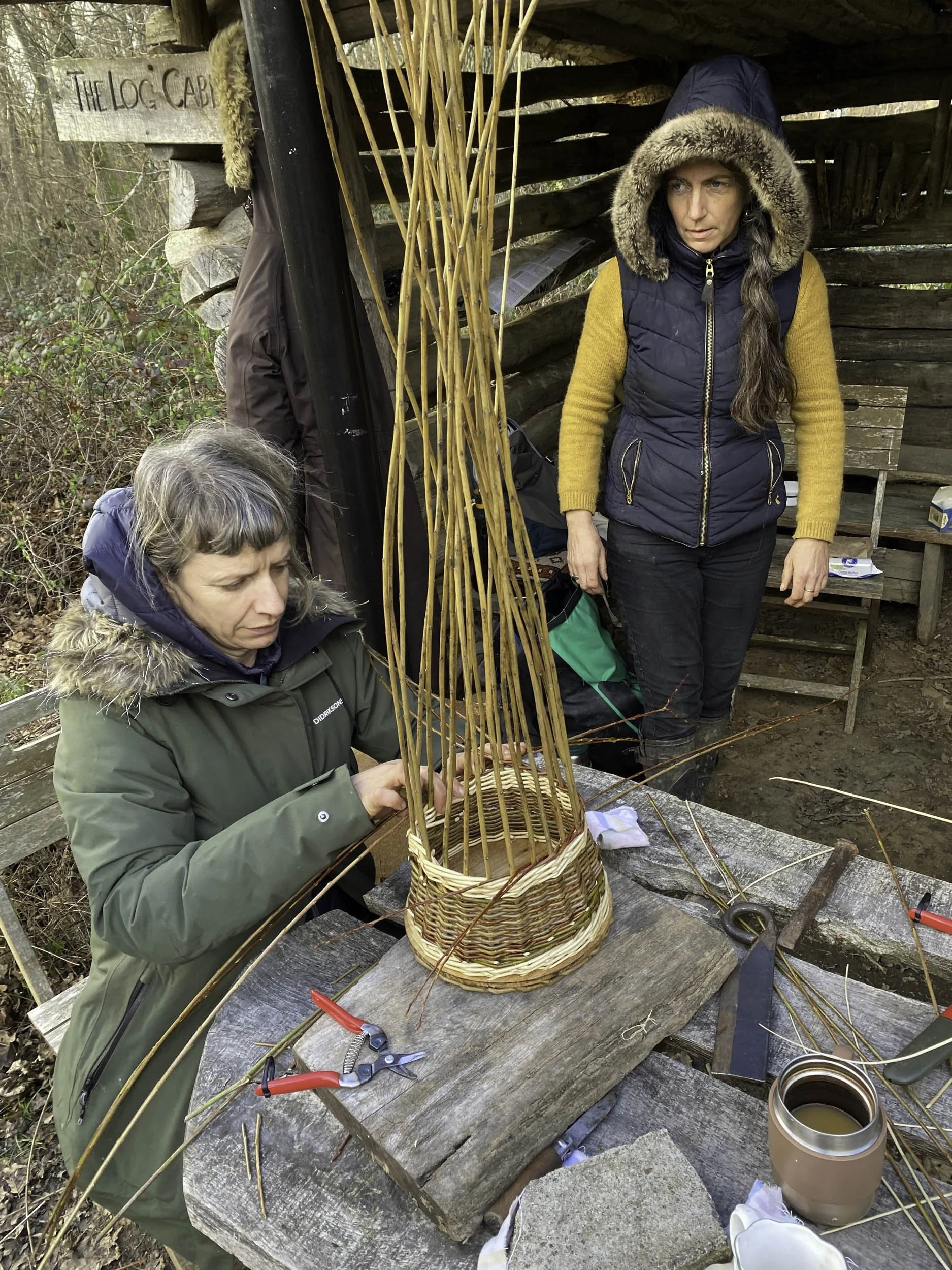 Two women outdoors working on wicker basket weaving at a rustic wooden table. One woman is sitting, the other is standing with a hooded jacket. They are surrounded by natural woodland.