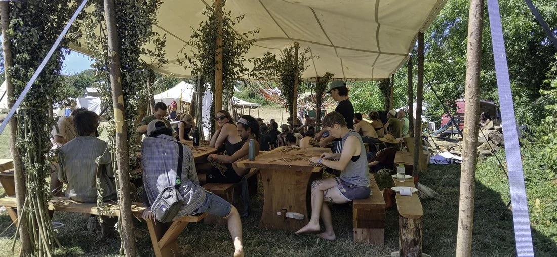 People sitting at wooden picnic tables under a canopy in a grassy outdoor area surrounded by trees and tents.