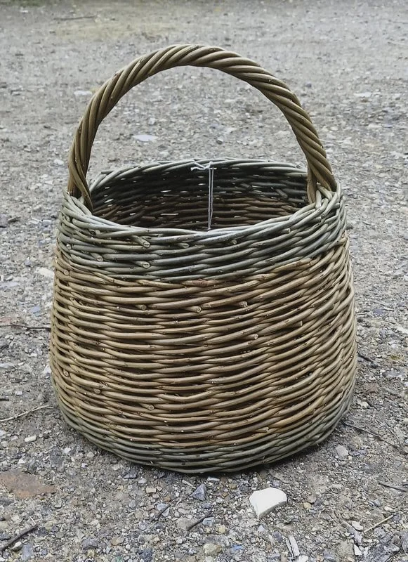 A woven wicker basket with a handle, sitting on gravel or dirt ground.
