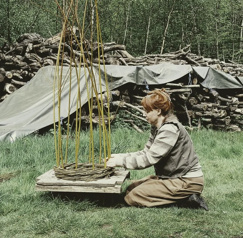 A woman with short red hair kneeling on grass, assembling a wicker basket with tall, thin wooden poles outside in a wooded area.