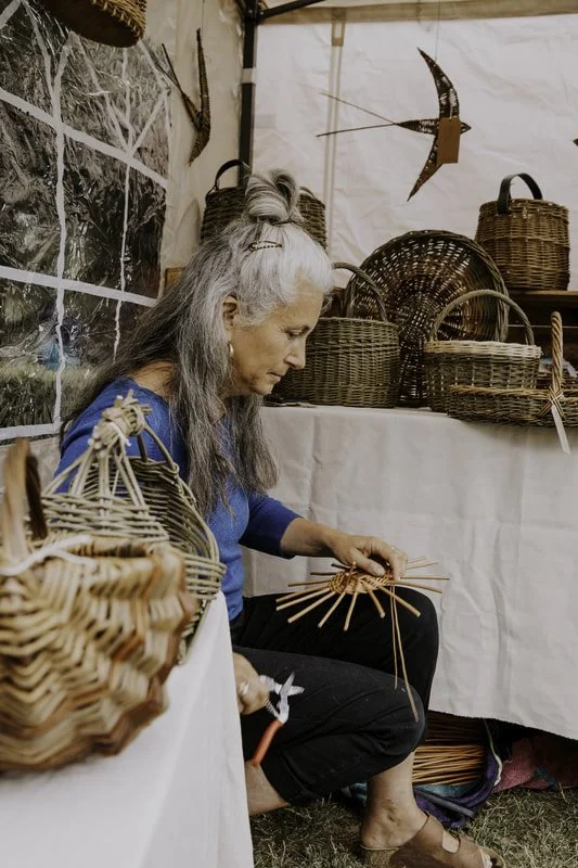 A woman with gray hair sitting on the ground and weaving a basket using natural material. She is wearing a blue shirt and black pants, surrounded by finished woven baskets and craft supplies in a craft booth or market stall.