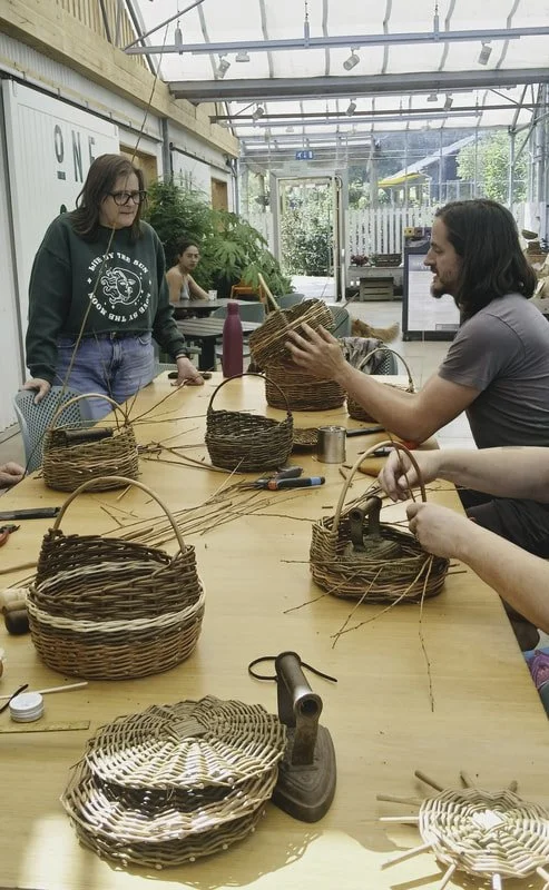 People working on basket weaving project at a long wooden table in a bright, enclosed workspace.