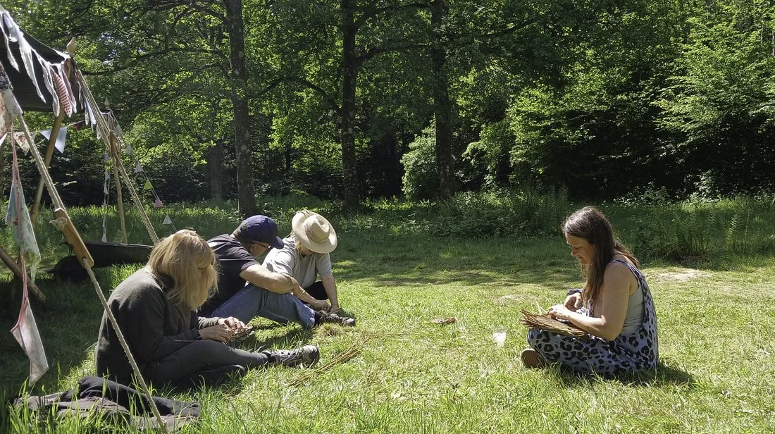 Four children sitting on grass in a forest, engaged in arts and crafts, with a young woman sitting facing them also working on arts and crafts.