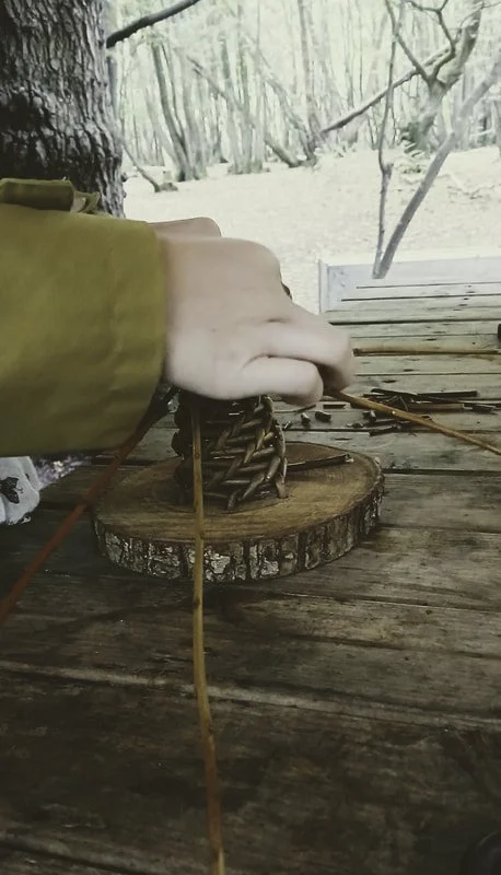 A person wearing a yellow jacket is tying a small wooden or metal chain onto a tree trunk with a wooden table surface in the foreground and a snowy forest scene in the background.