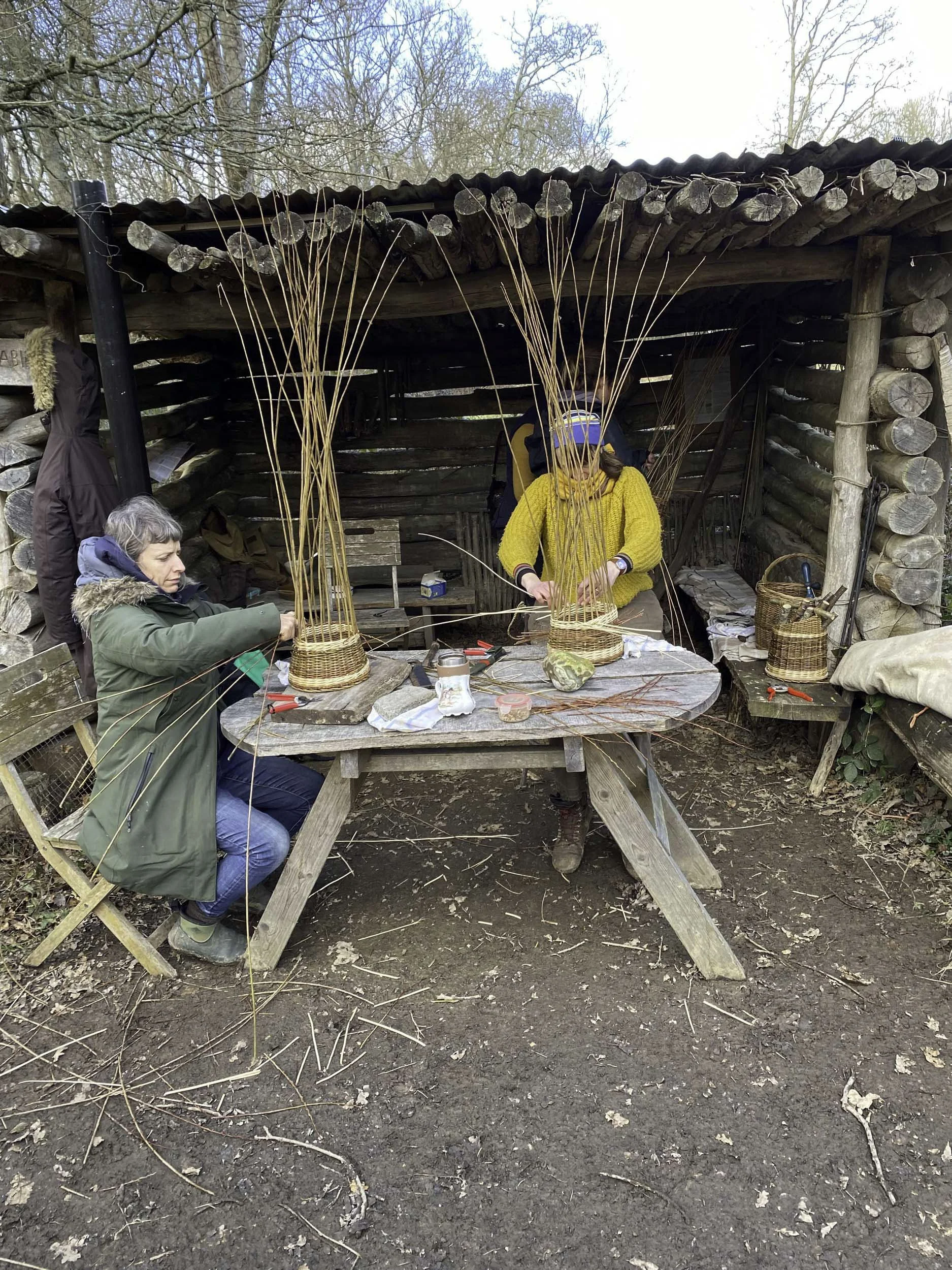 Two people working on a craft project outdoors at a rustic wooden table, constructing baskets with long, thin wicker or reed materials. A small shed or shelter is behind them, made of logs, with trees in the background.