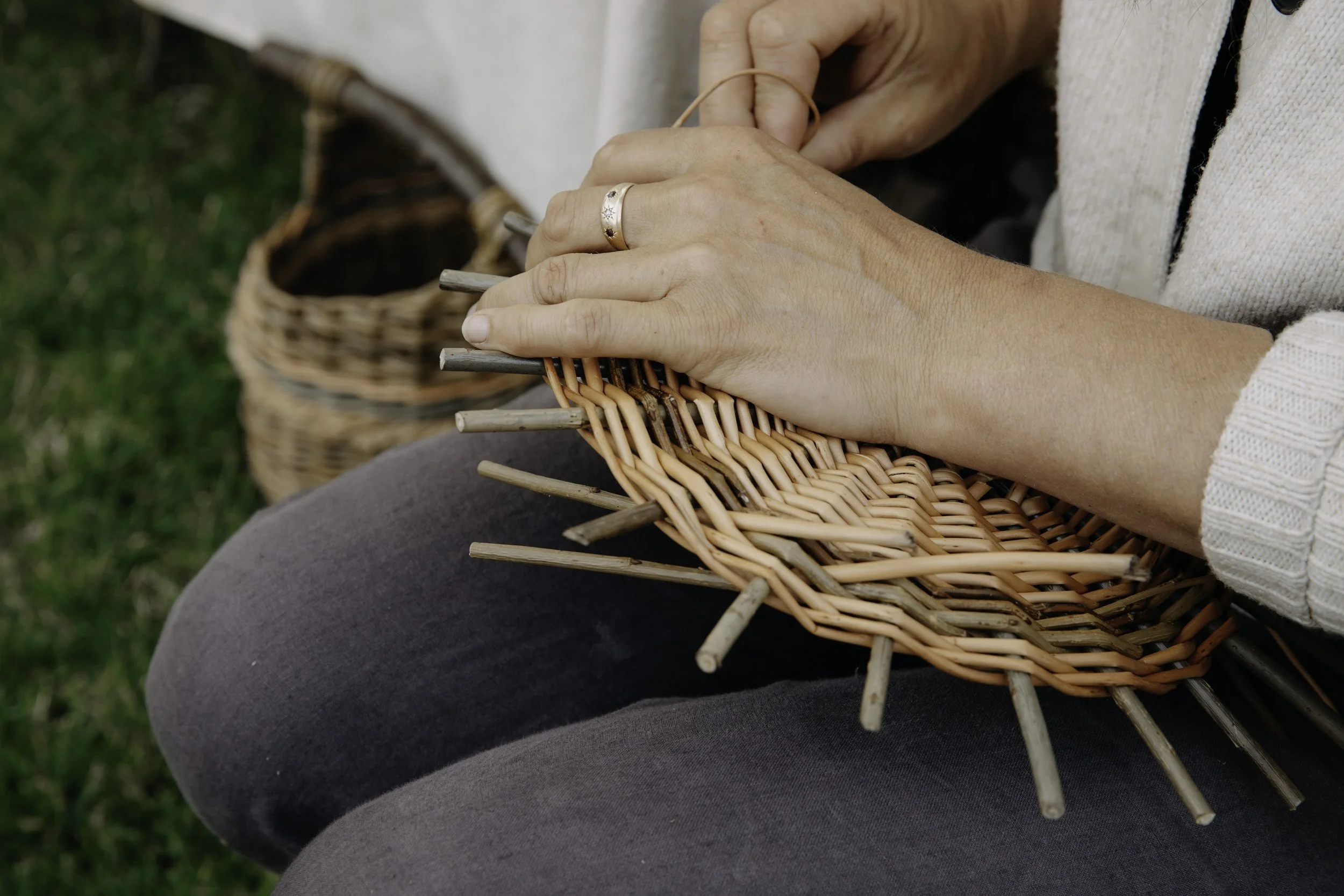 Close-up of hands weaving a wicker basket outdoors, with a basket in the background on green grass.