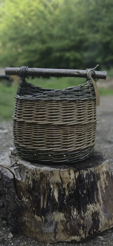 A wicker basket hanging from a wooden stick resting on a tree stump outdoors, with greenery in the background.