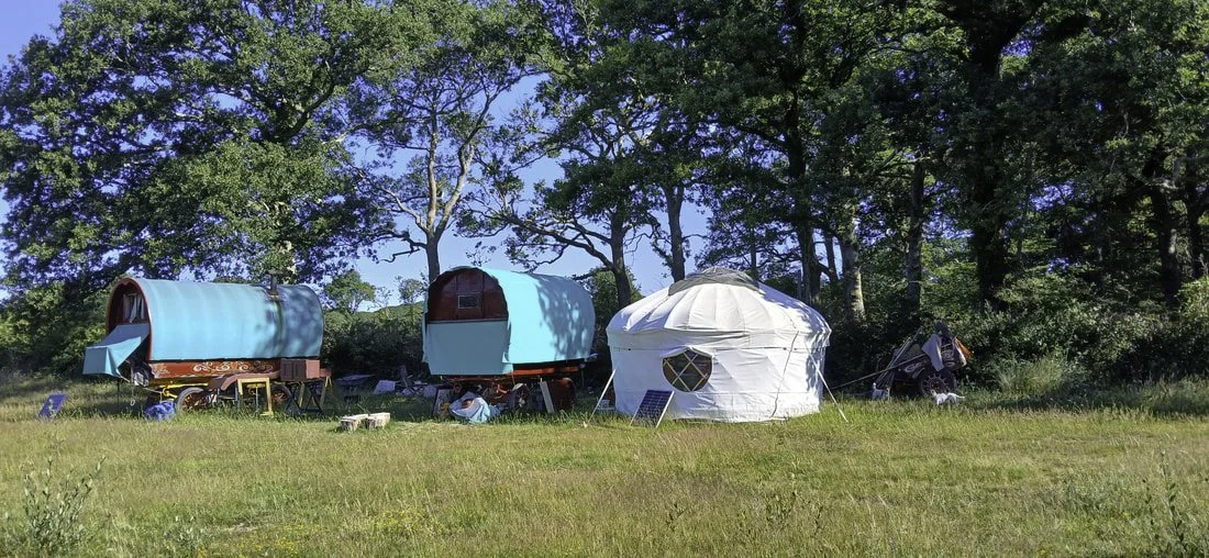Two colorful vintage caravans and a white yurt set up on a grassy field, surrounded by trees under a blue sky.