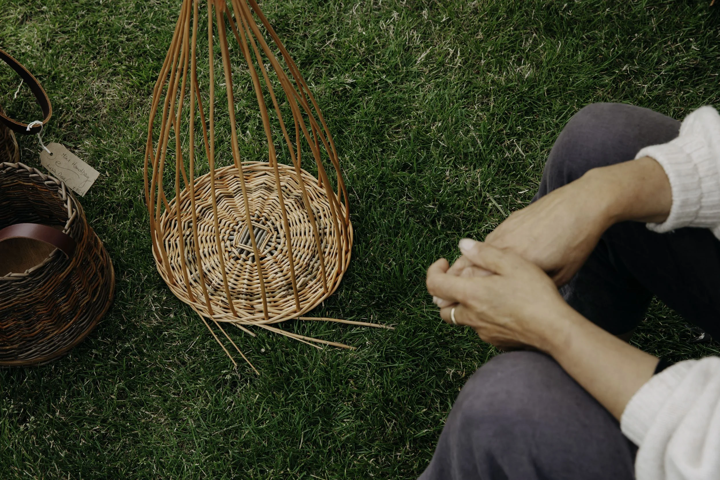 Lisa Dear sitting on grass, holding hands, surrounded by wicker baskets and baskets with tags, in an outdoor setting.