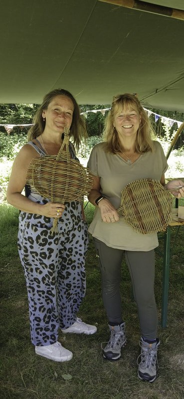 Two women standing outdoors under a tent, holding woven baskets and smiling.